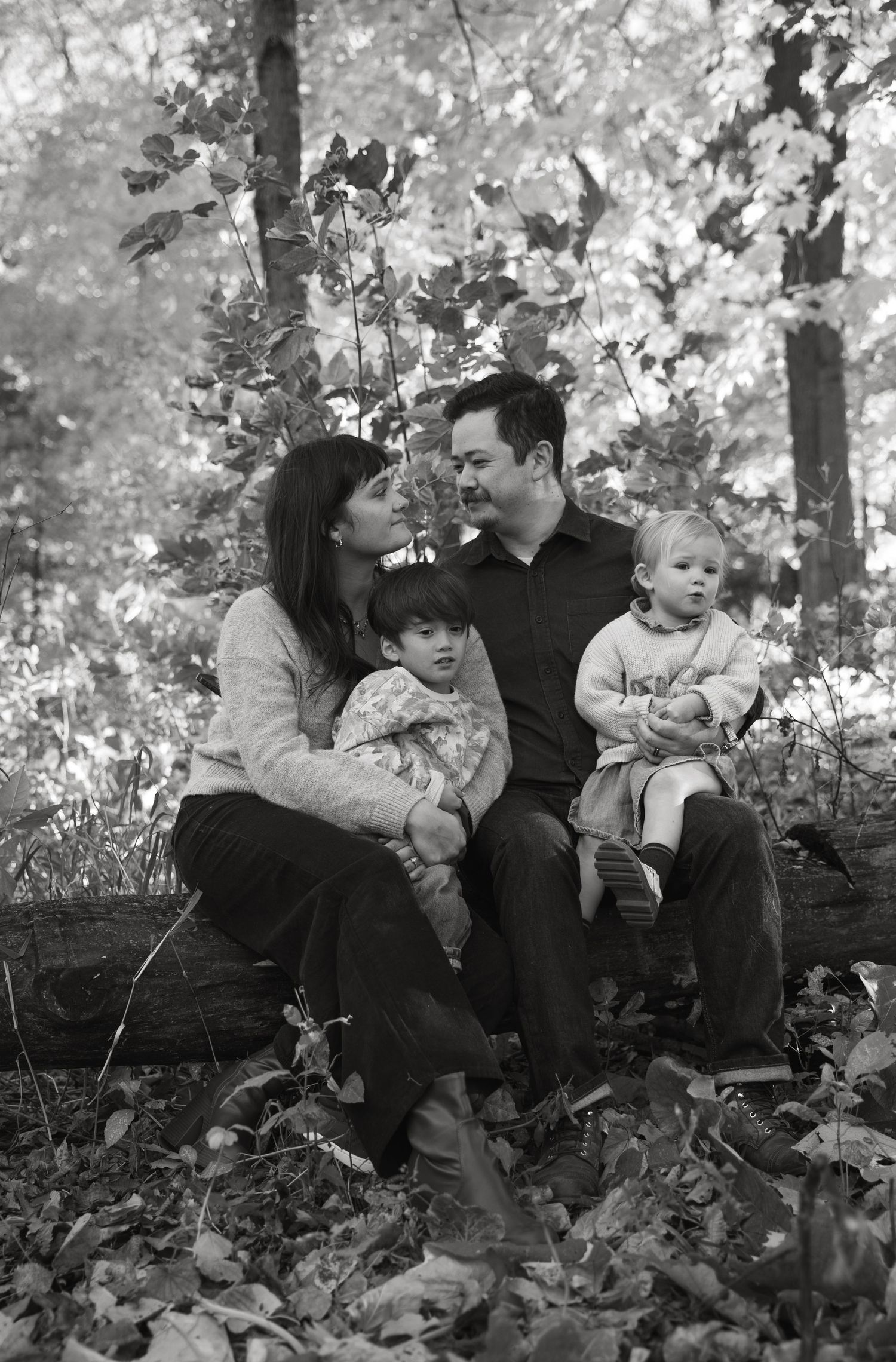 A smiling family of four sits close together on a log among fallen autumn leaves in a woodland, in a black and white portrait.