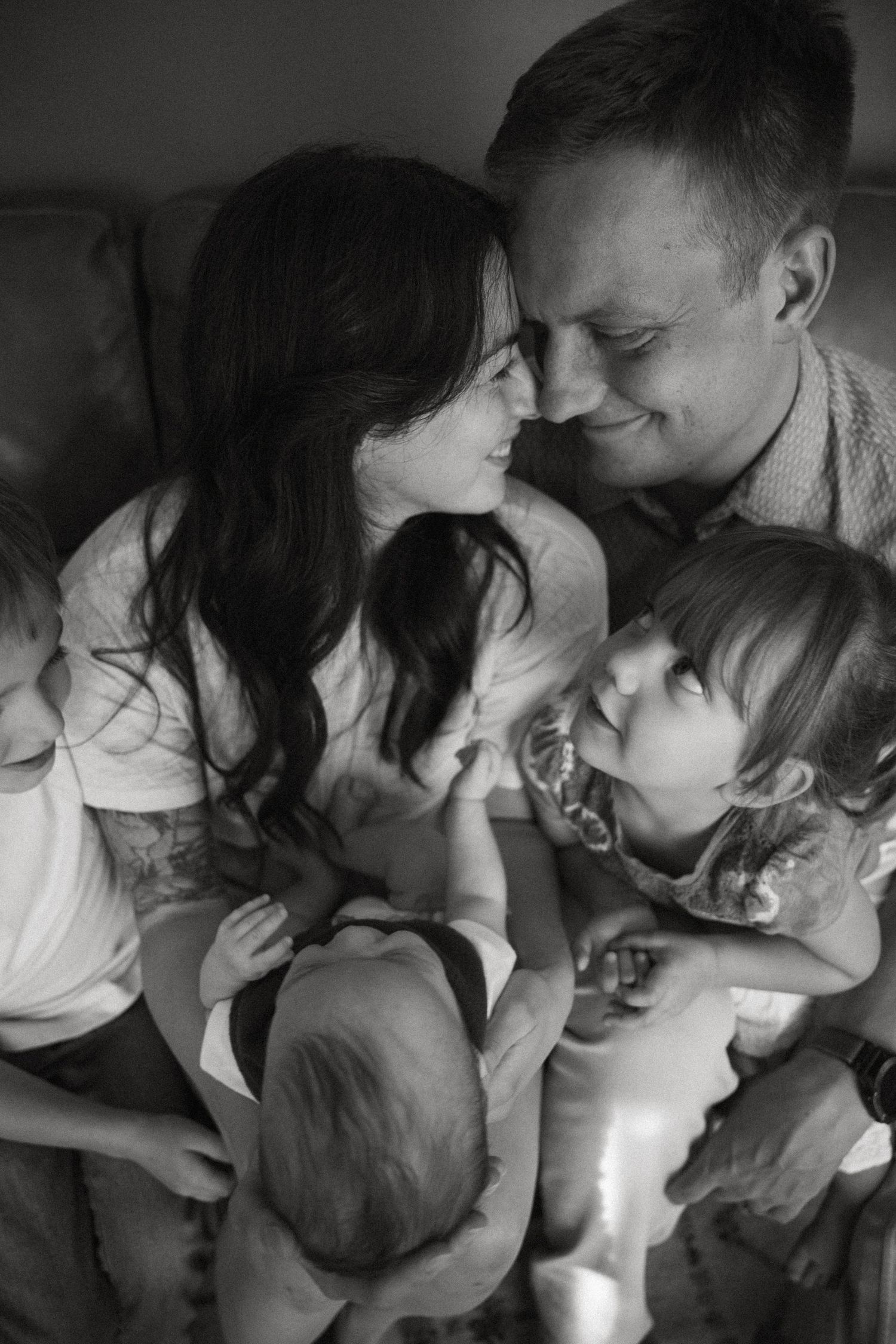 A black and white close-up shows two parents smiling nose to nose, surrounded by their three young children in a cozy embrace.