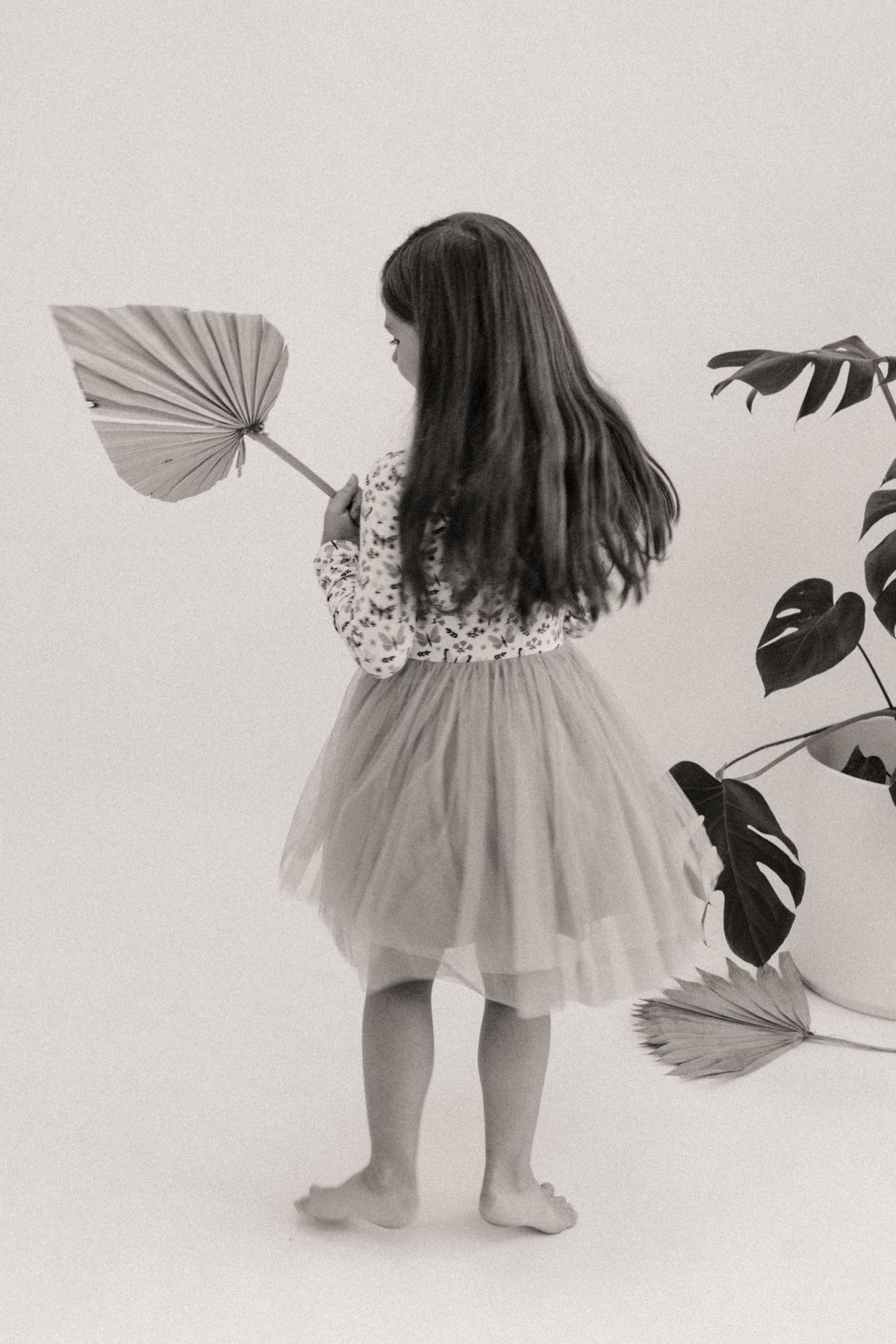 Black and white photo of a little girl with long hair in a tulle skirt holding a dried palm leaf fan beside a monstera plant.