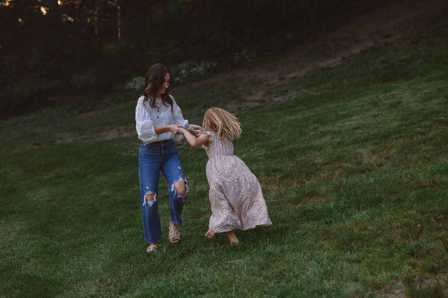 A woman in jeans holds hands with a young girl in a floral dress as they spin and play on a grassy hillside.