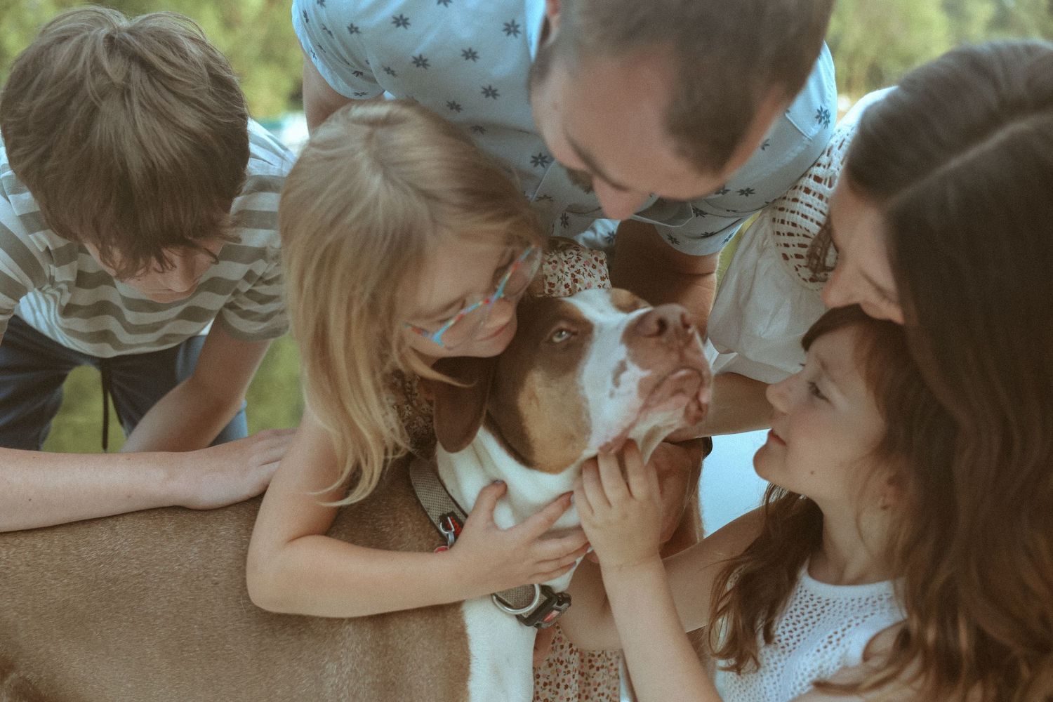 A family with three children and two adults gathers close to pet and kiss a brown and white dog outdoors.