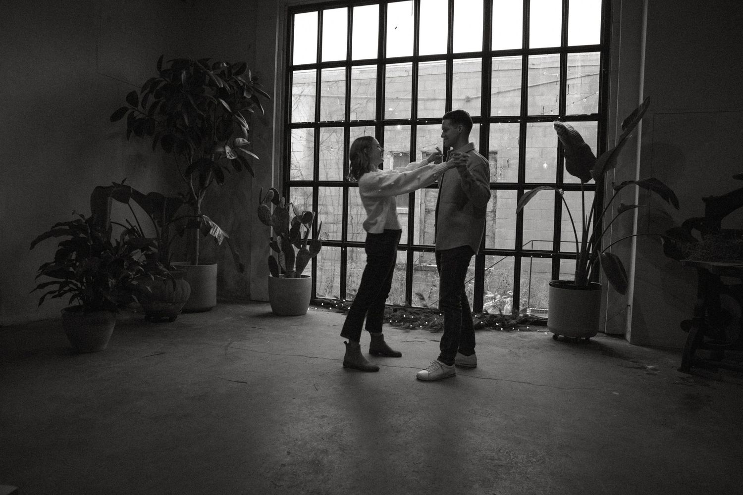 A black and white photo of a couple dancing in a dim room with large industrial windows and potted plants.