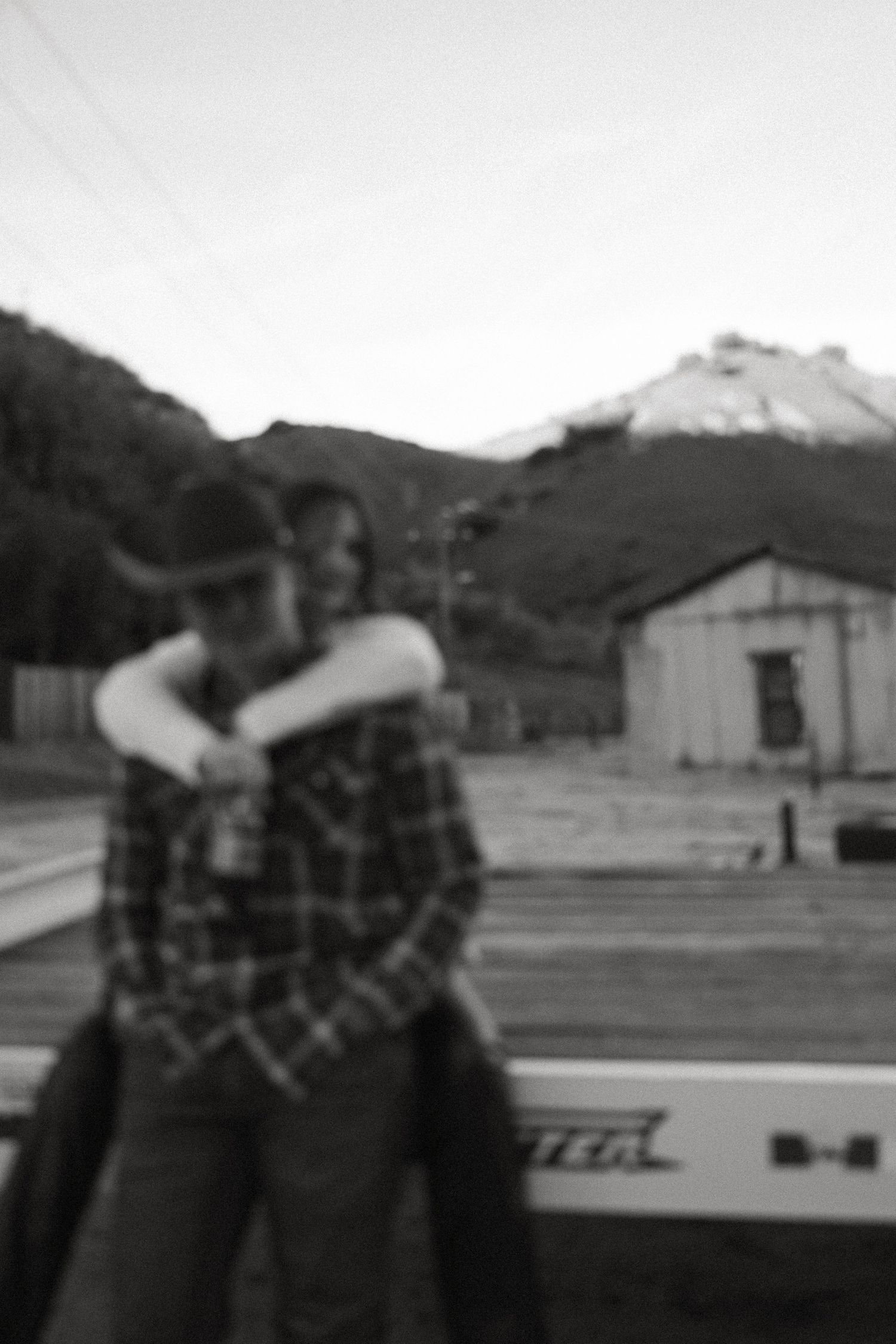 Blurry black and white photo of a person in a cowboy hat and plaid shirt being hugged from behind, with a barn and snowy mountain behind.