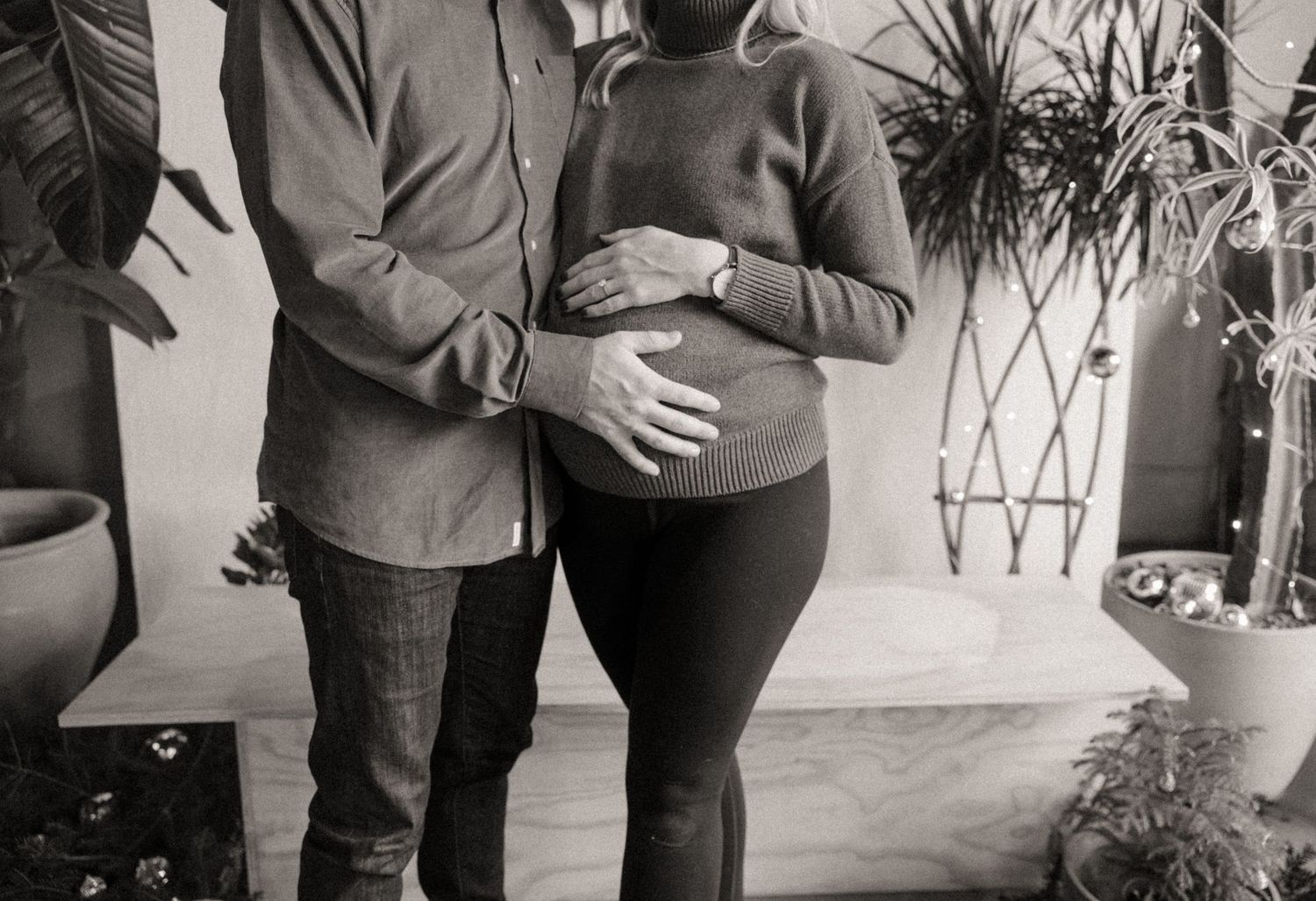Black and white photo of an expecting couple standing close together, hands cradling the pregnant belly, surrounded by green plants.