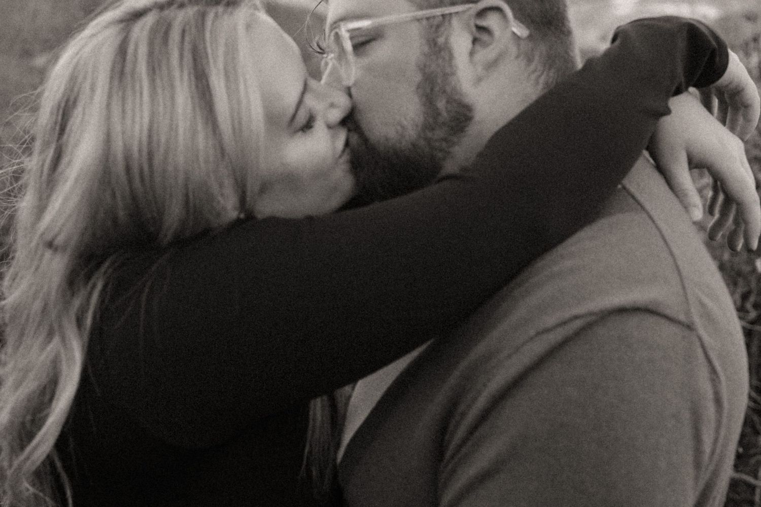 Black and white close-up of a couple sharing a kiss outdoors, the man bearded with glasses and the woman embracing him.