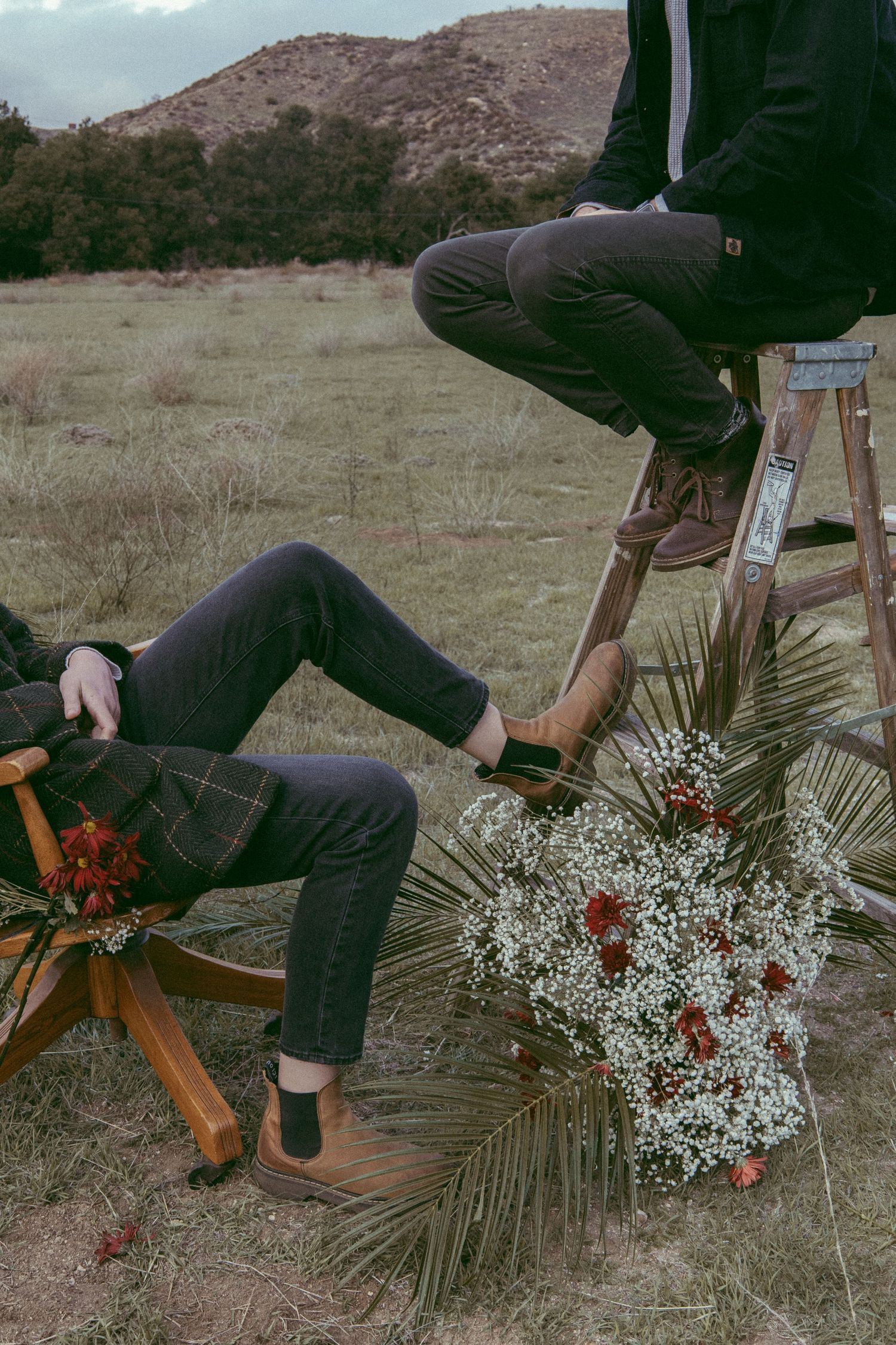 Two people in boots sit on a wooden chair and ladder in a dry field, with a bouquet of baby's breath, palm leaves and red blooms.
