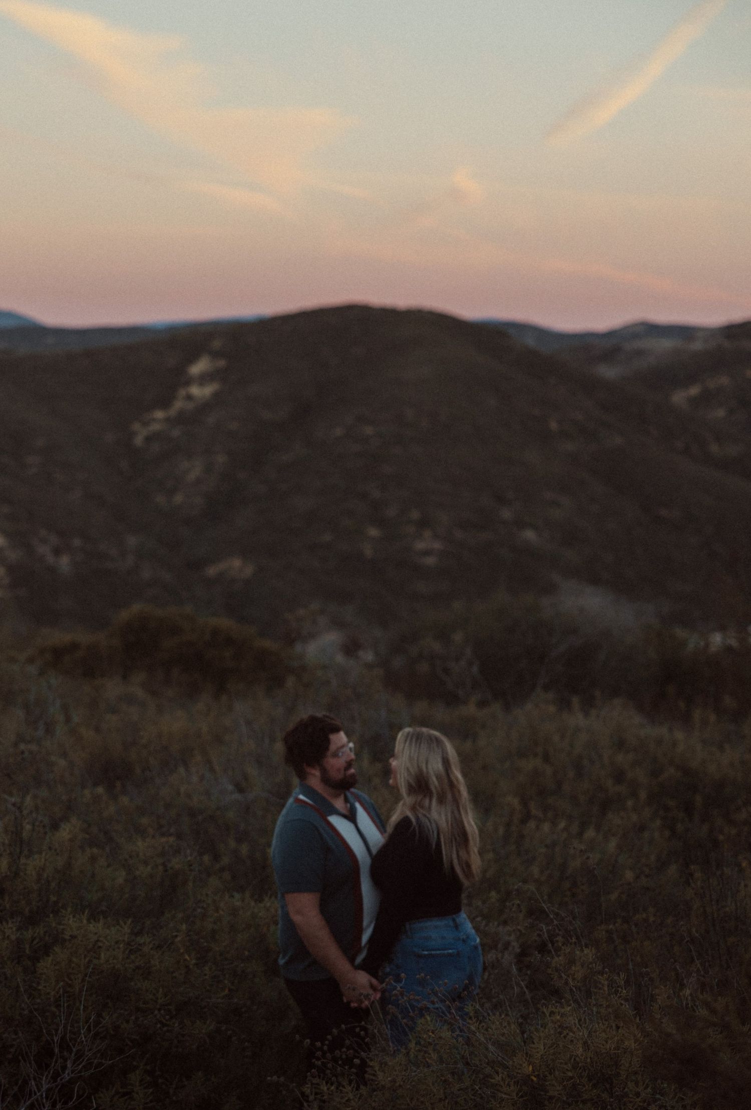 Couple embraces on a hillside covered in shrubs as a pink sunset glows over dark mountains in the background.