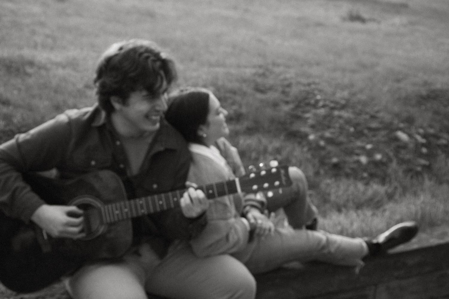 Black and white photo of a smiling man playing acoustic guitar while a woman sits beside him on a grassy hillside.