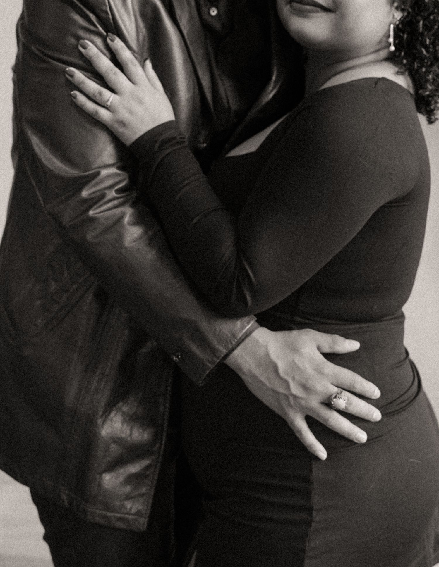 Black and white close-up of a couple embracing, the woman's hands showing rings as the man rests a hand on her waist.