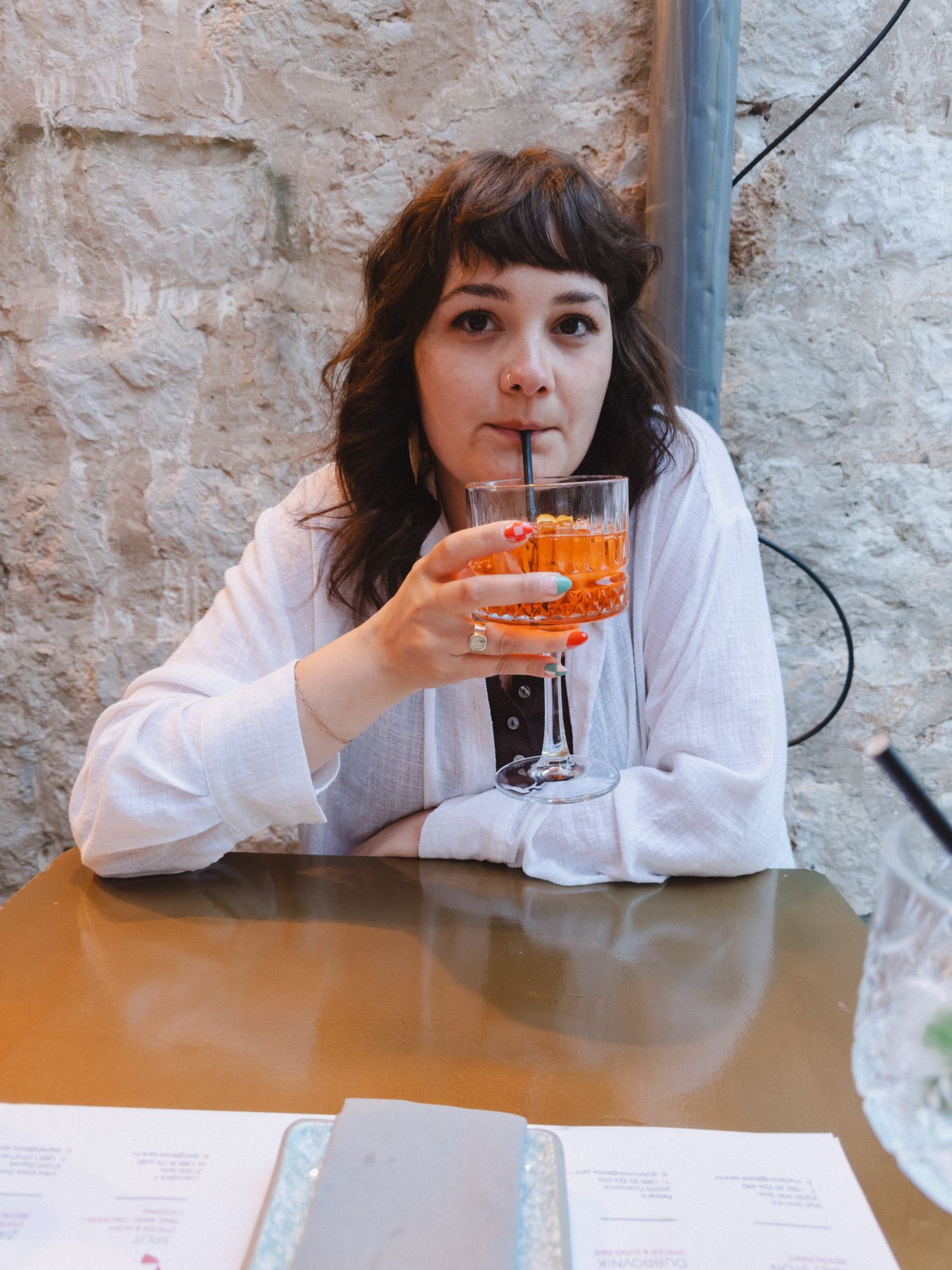 A woman with dark wavy hair and bangs sips an orange spritz through a straw at a restaurant table by a stone wall.