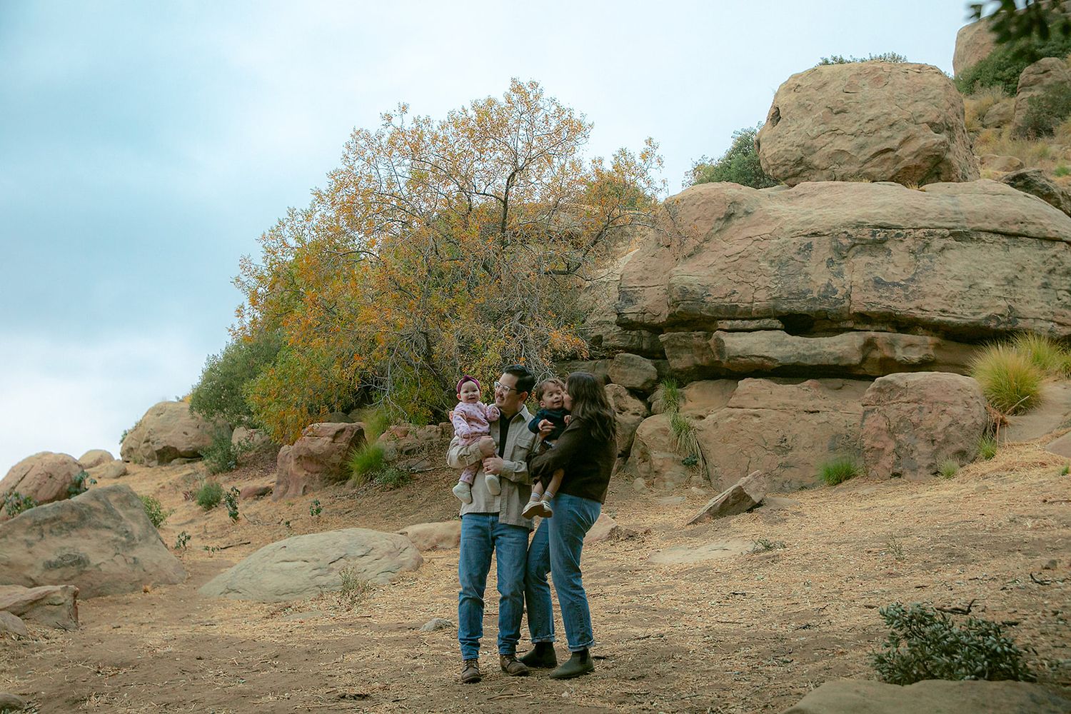 Family of three stands among large boulders and autumn trees on a rocky desert trail, holding a small child.
