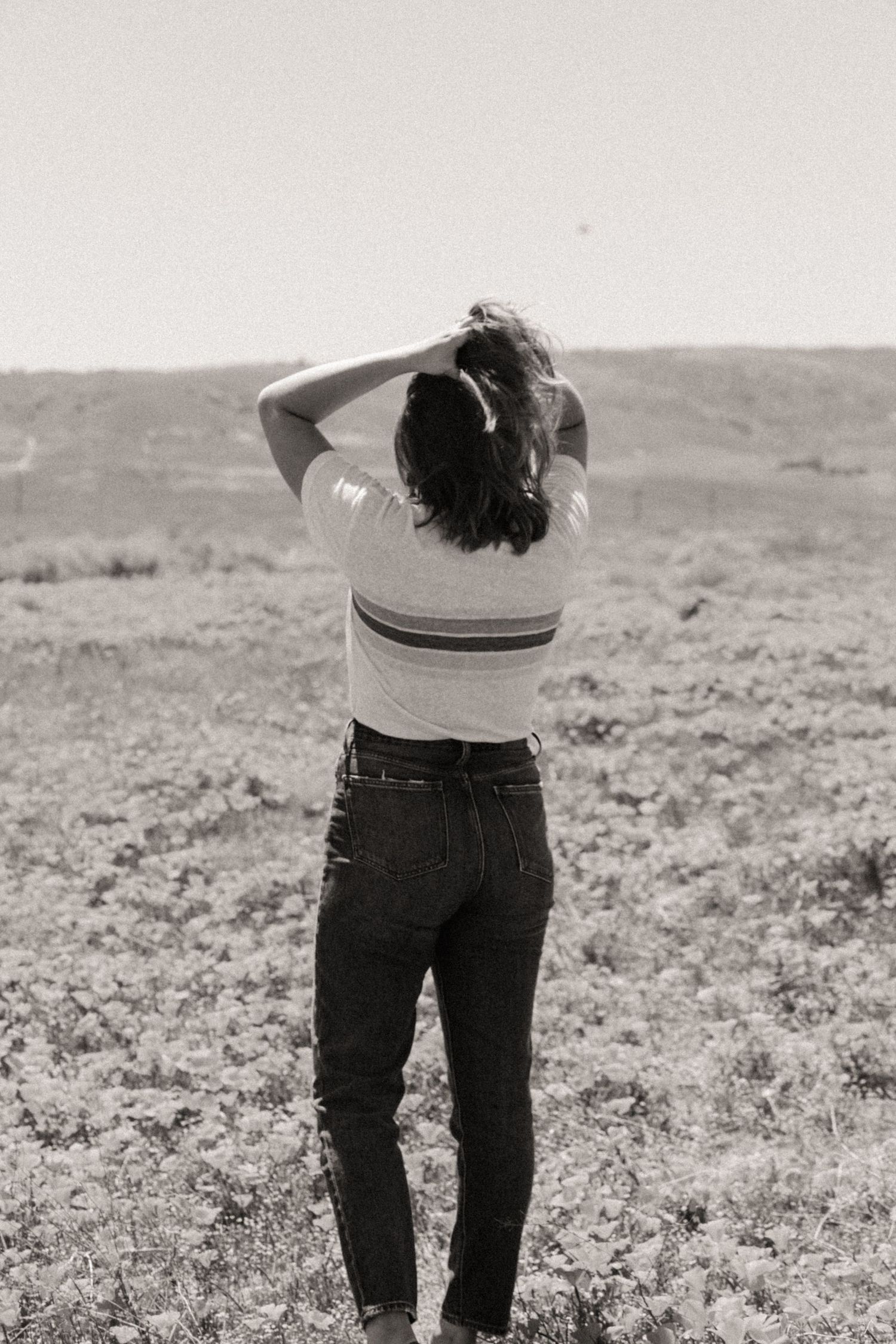 Black and white photo of a woman in a striped tee and jeans standing in a field with her hands in her hair.