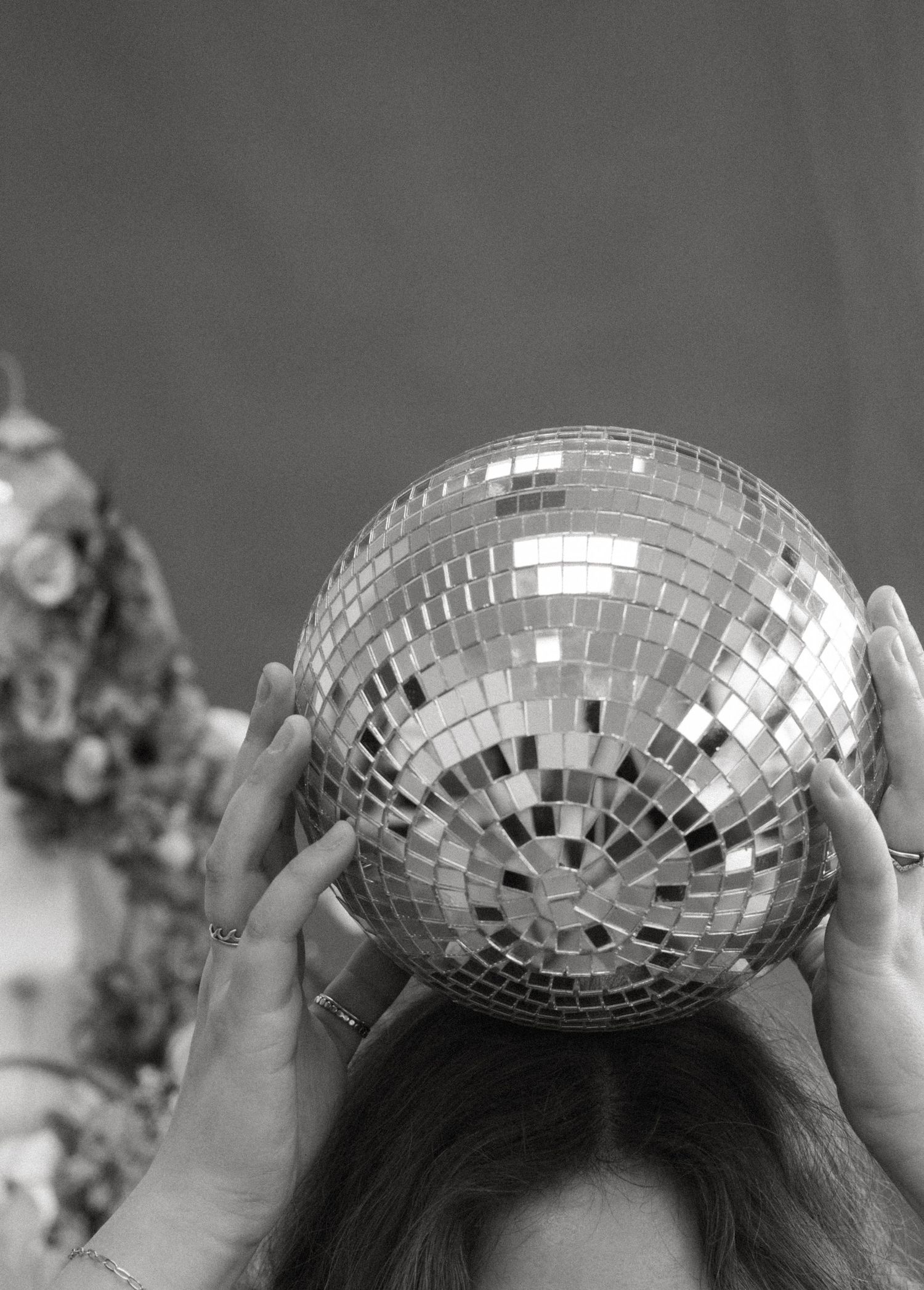 Black and white close-up of hands holding a mirrored disco ball above a woman's head, with flowers softly blurred behind.