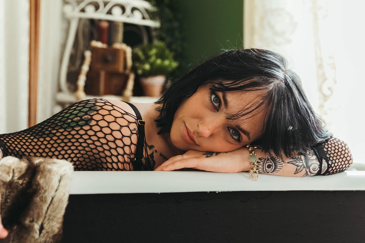 Woman with dark hair and tattoos in fishnet top leaning on bathtub edge in vintage bathroom setting.