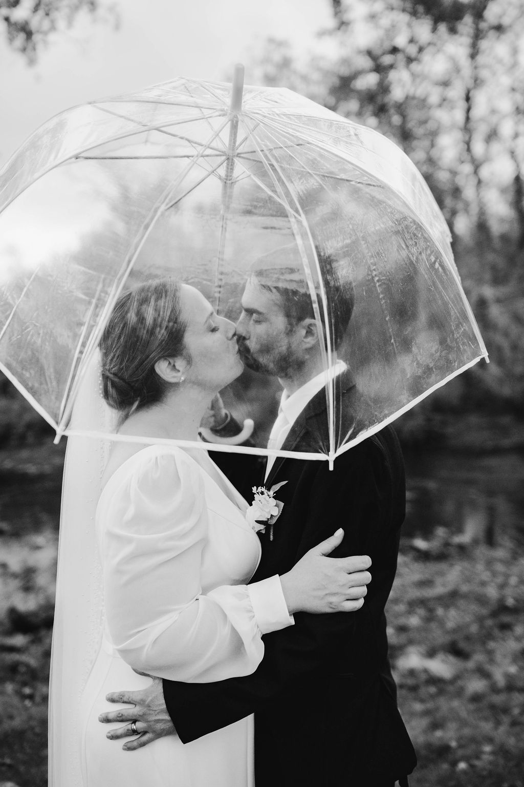 Black and white photo of a bride and groom kissing under a clear bubble umbrella outdoors on their wedding day.