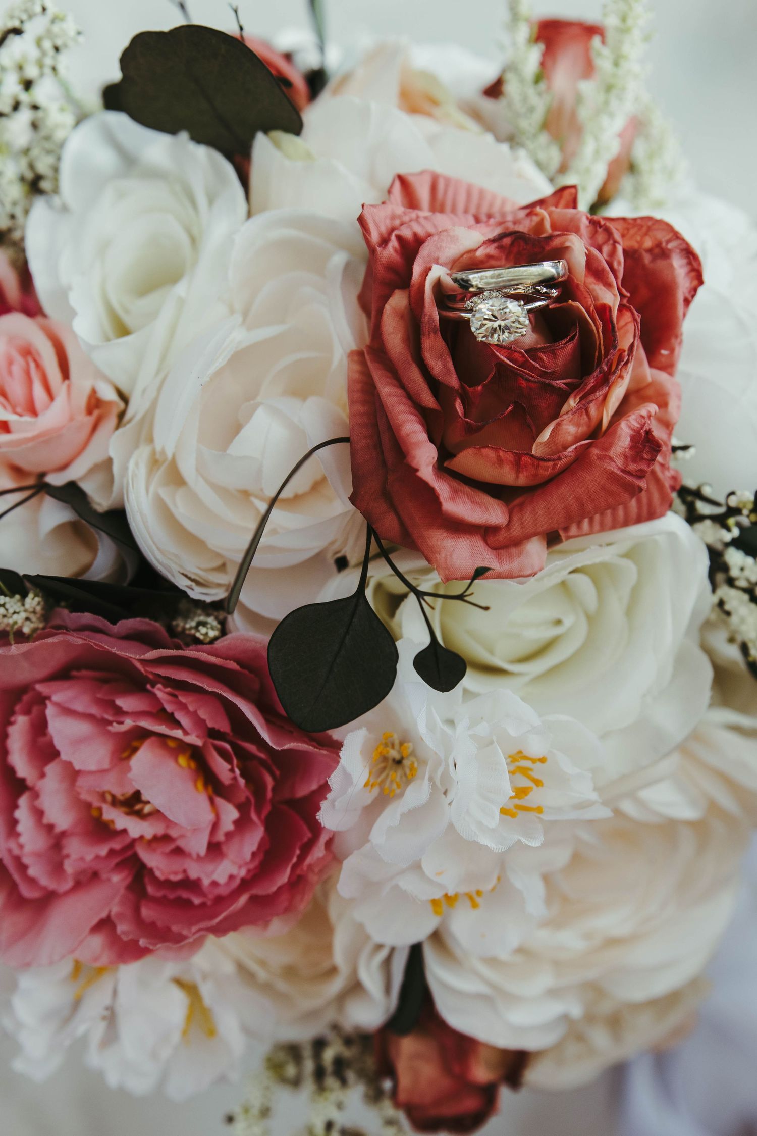 Elegant wedding bouquet with red, pink, and white roses featuring a diamond engagement ring nestled in a blooming flower.