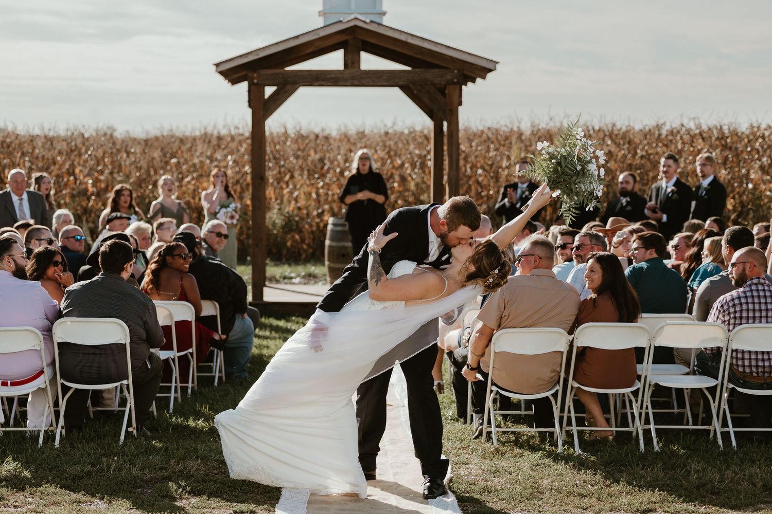 Bride and groom sharing a romantic dip kiss at an outdoor wedding ceremony beside a cornfield.