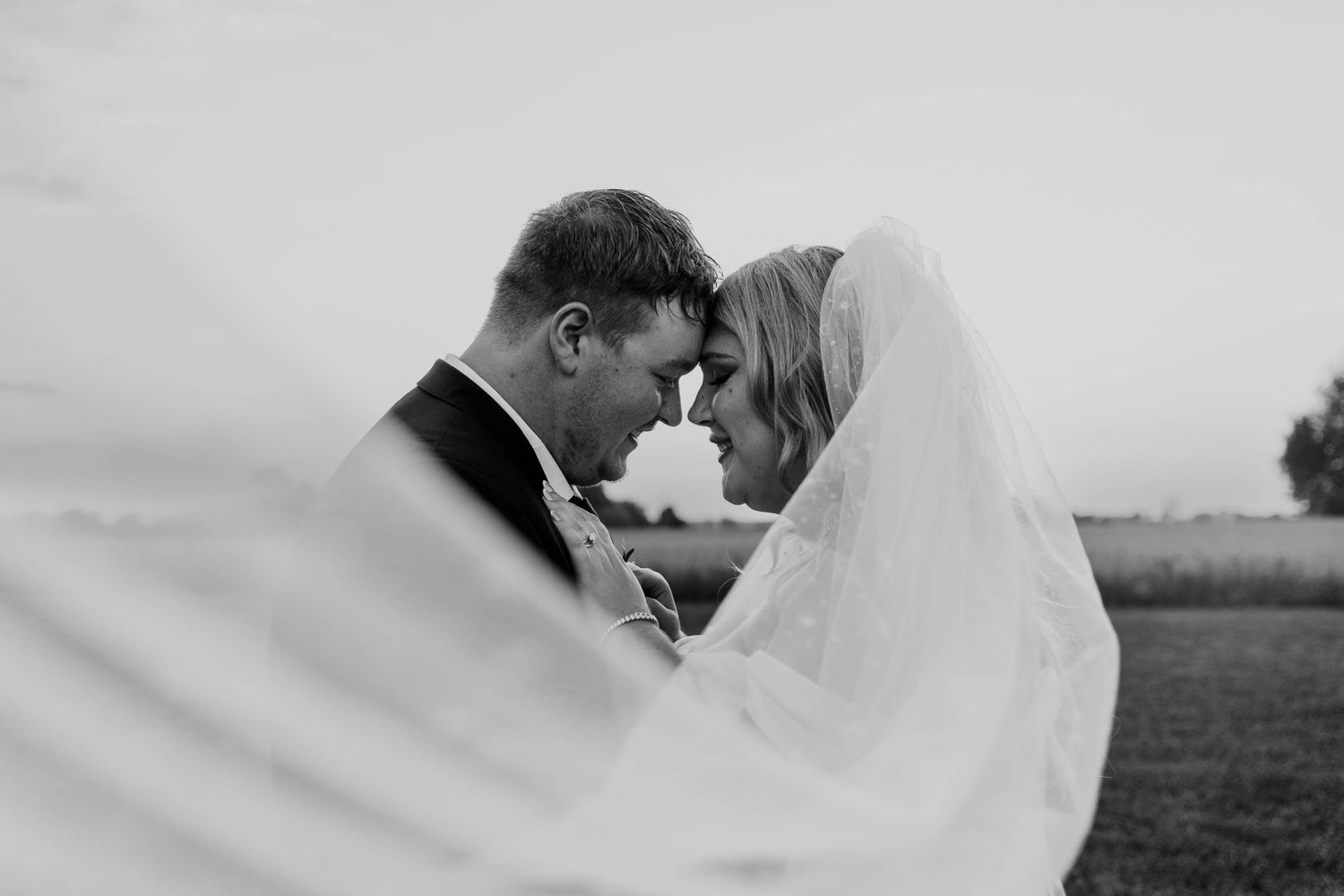Black and white photo of bride and groom touching foreheads, veil flowing around them in an outdoor field.