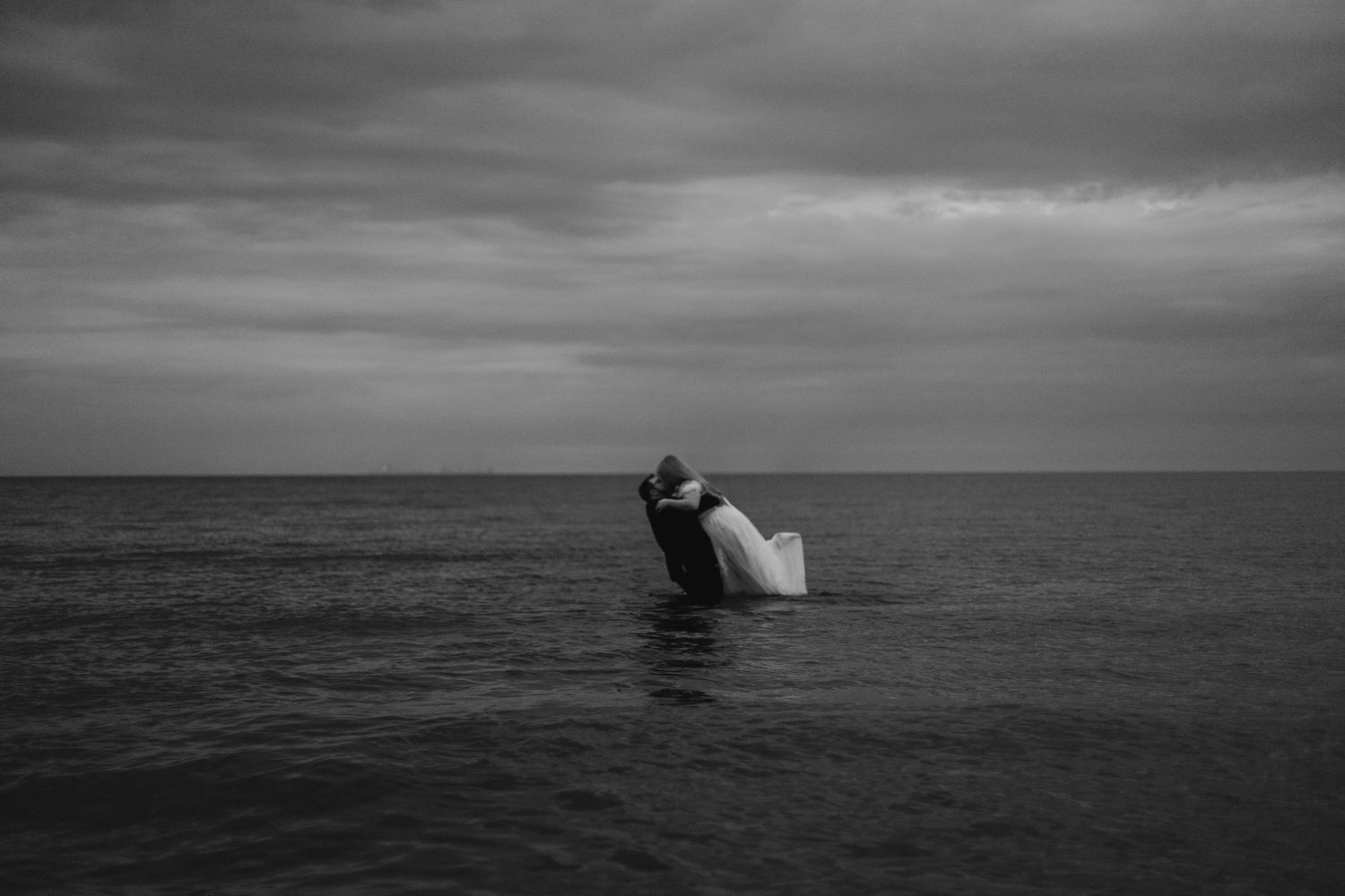 Black and white photo of a couple embracing in the ocean under a dramatic cloudy sky.
