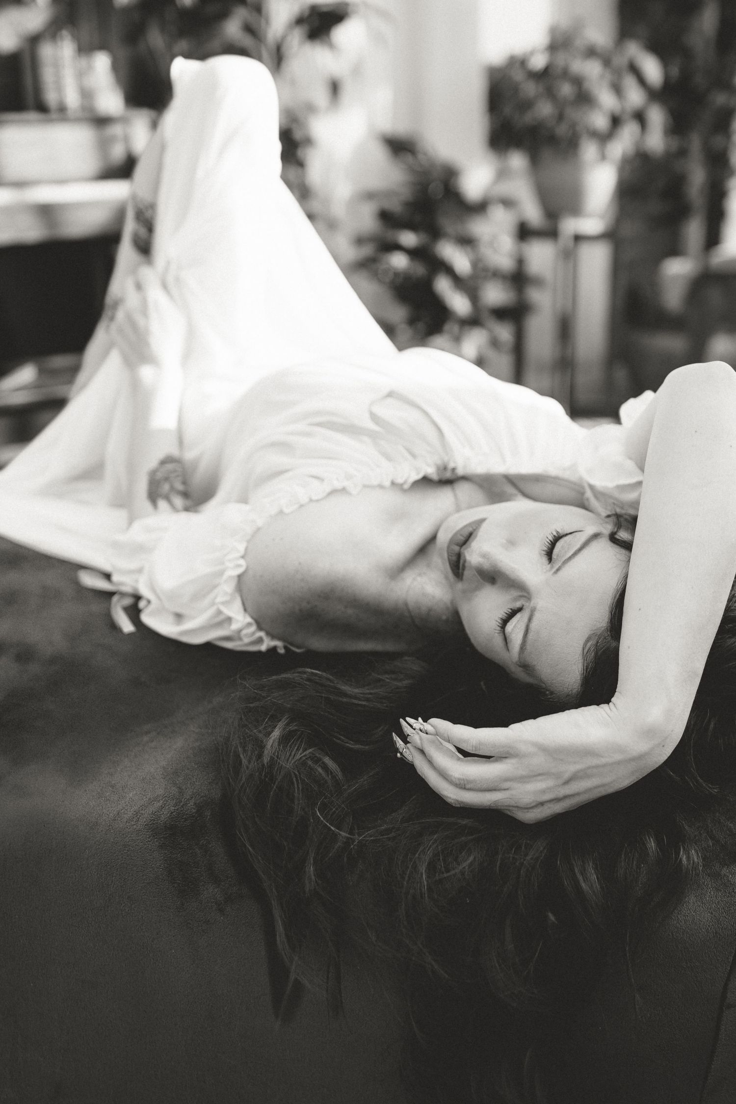 Black and white photo of a woman in white dress lying down with long hair spread out, eyes closed, indoors.