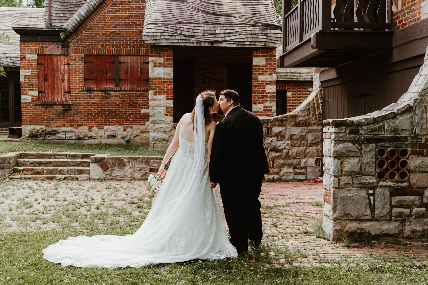 A bride and groom share a romantic kiss outdoors near a rustic brick building on their wedding day.