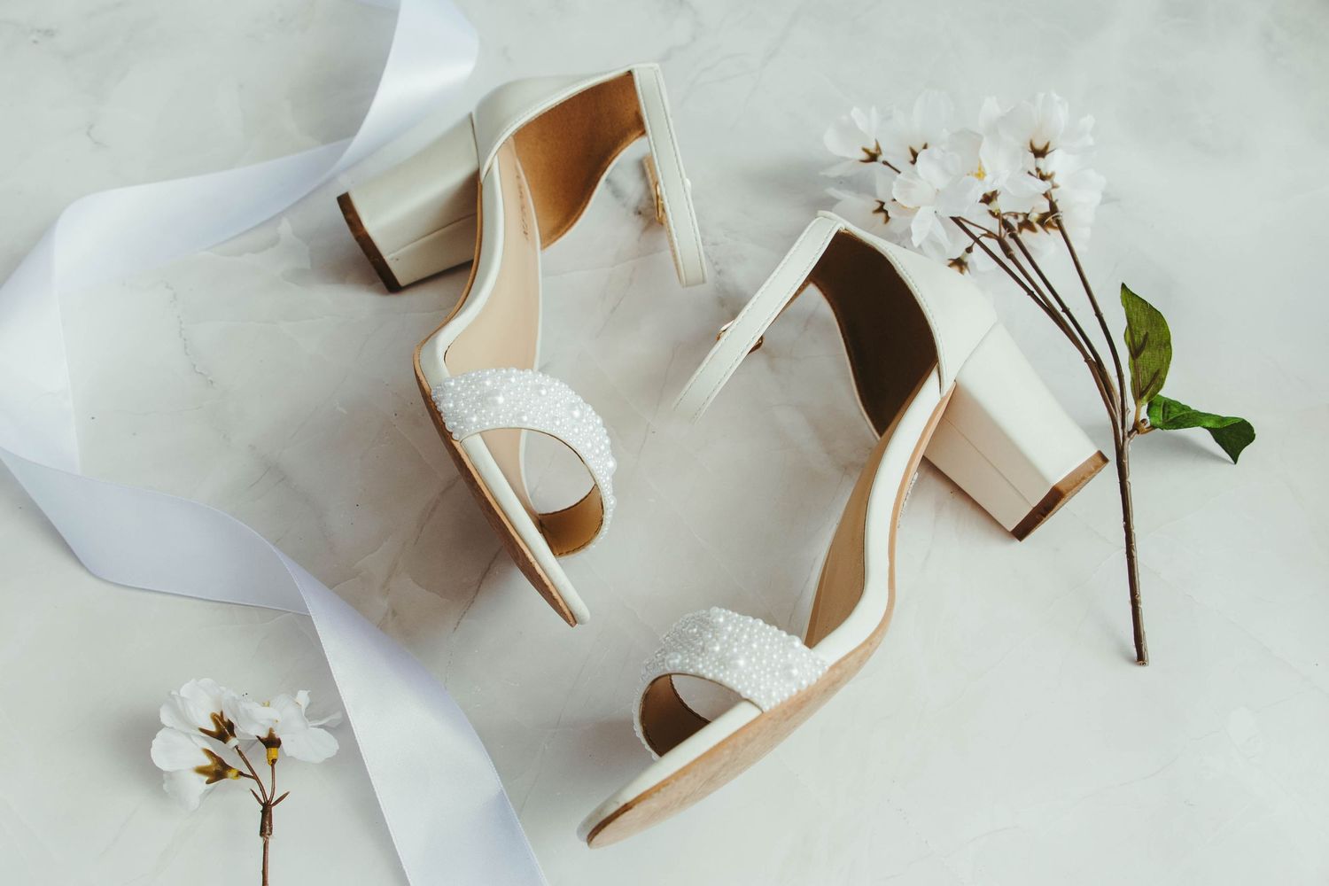 Elegant white bridal heels with pearl embellishments displayed with white cherry blossoms on marble.