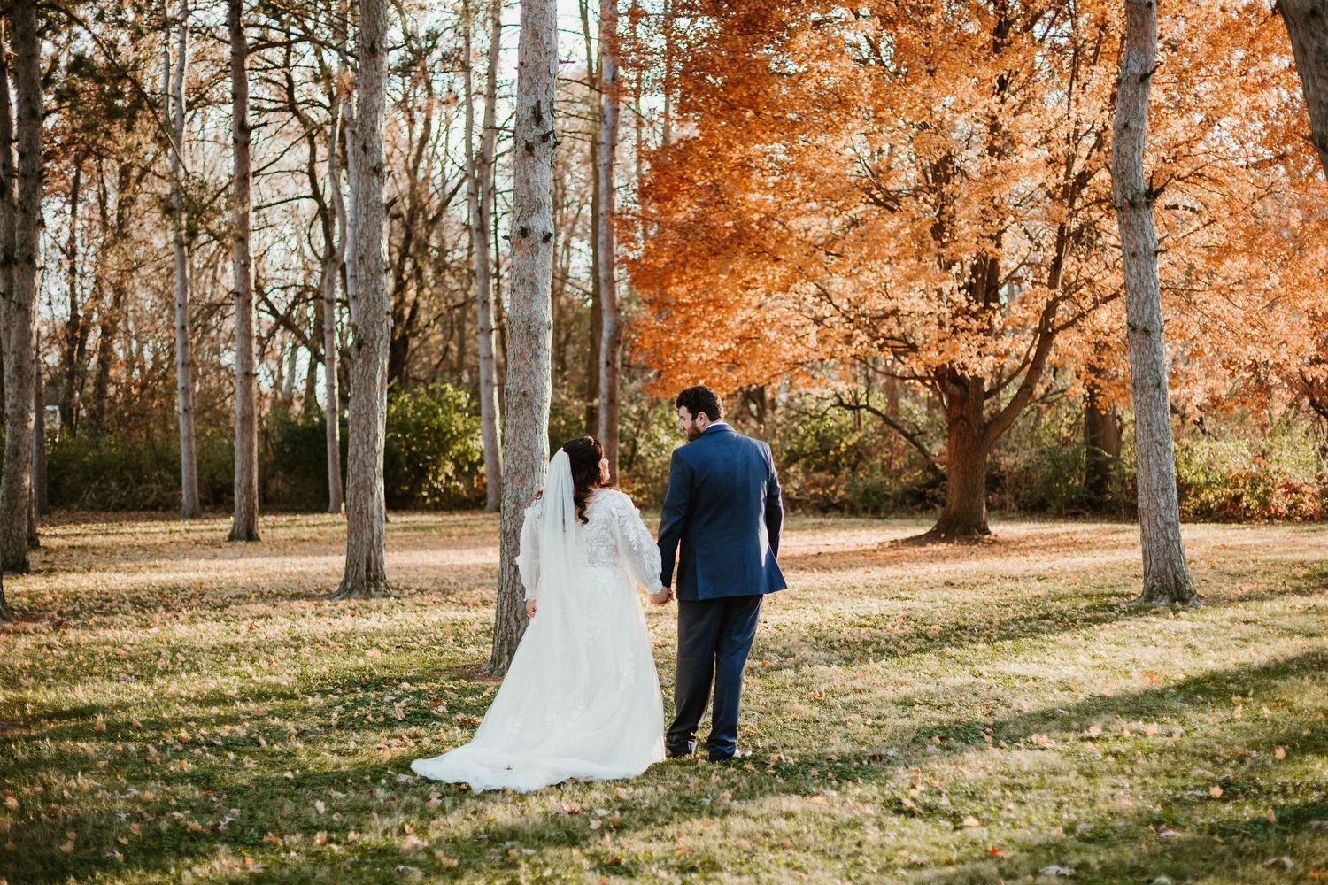 A bride and groom walk hand in hand through a scenic autumn park with vibrant orange foliage and tall trees.