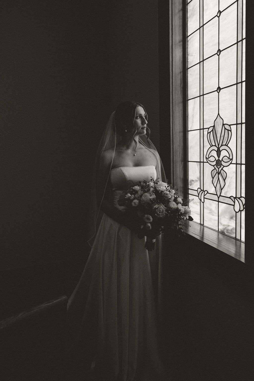 Black and white photo of a bride in a wedding gown holding a bouquet, standing by a stained glass window.