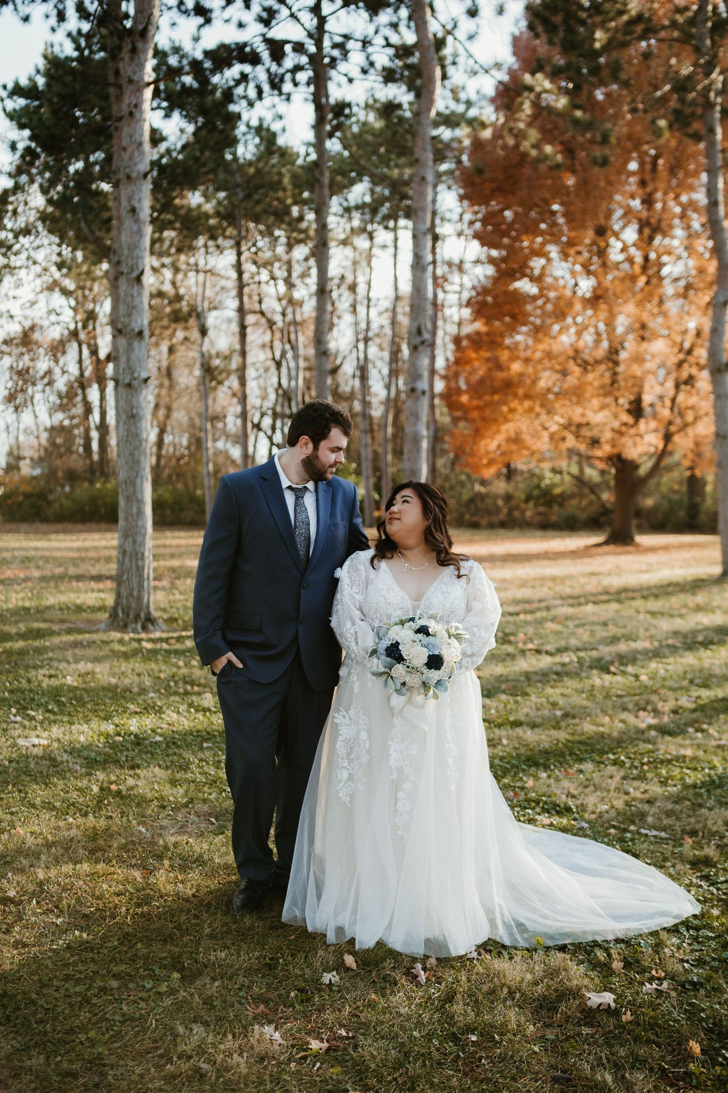 A bride and groom walk together in a park with autumn foliage, she holds a white bouquet in a lace gown.
