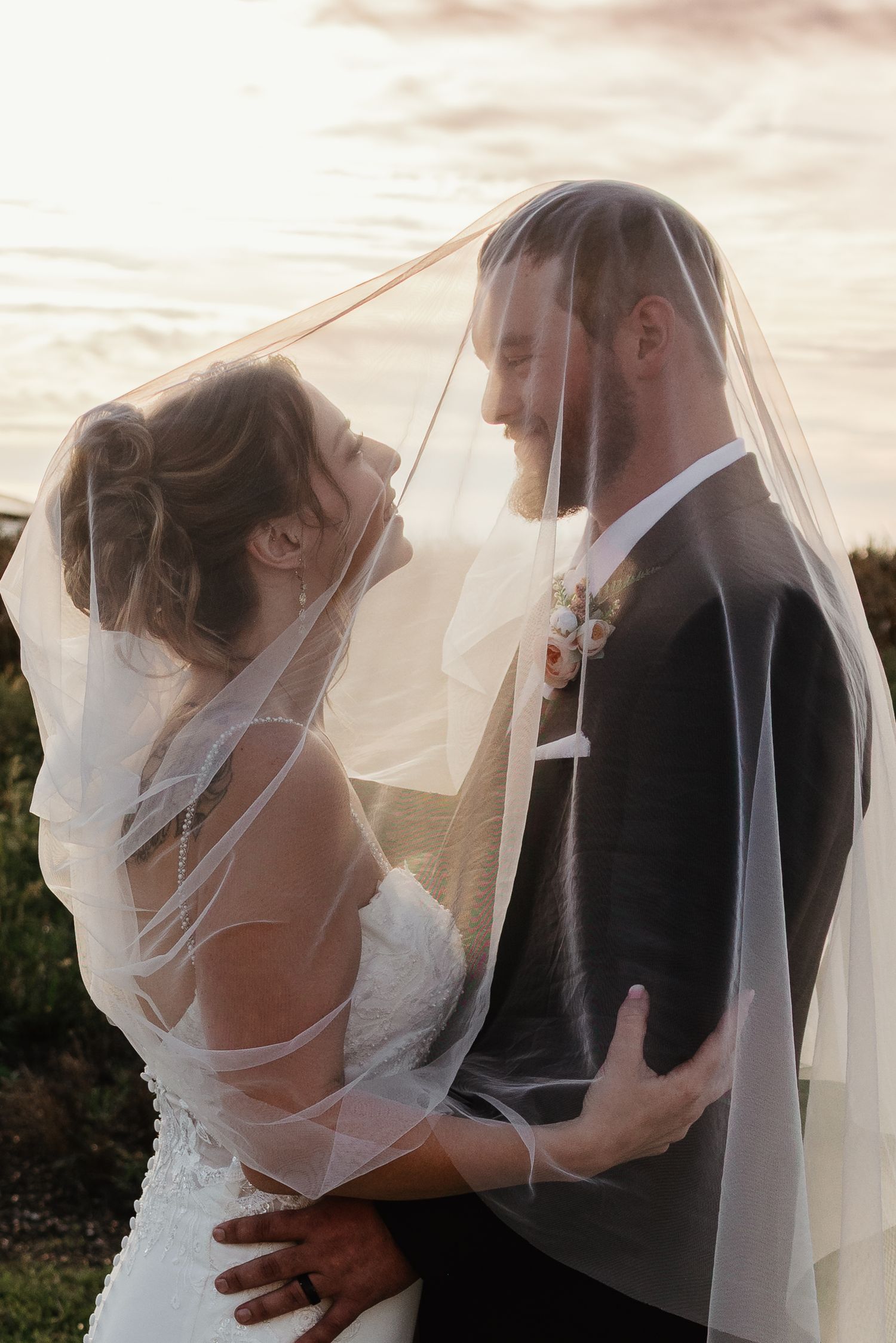 Romantic couple shares intimate moment under bridal veil at sunset by the ocean.