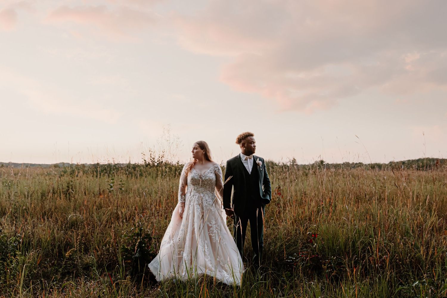 Bride and groom holding hands while walking through a golden meadow at sunset.