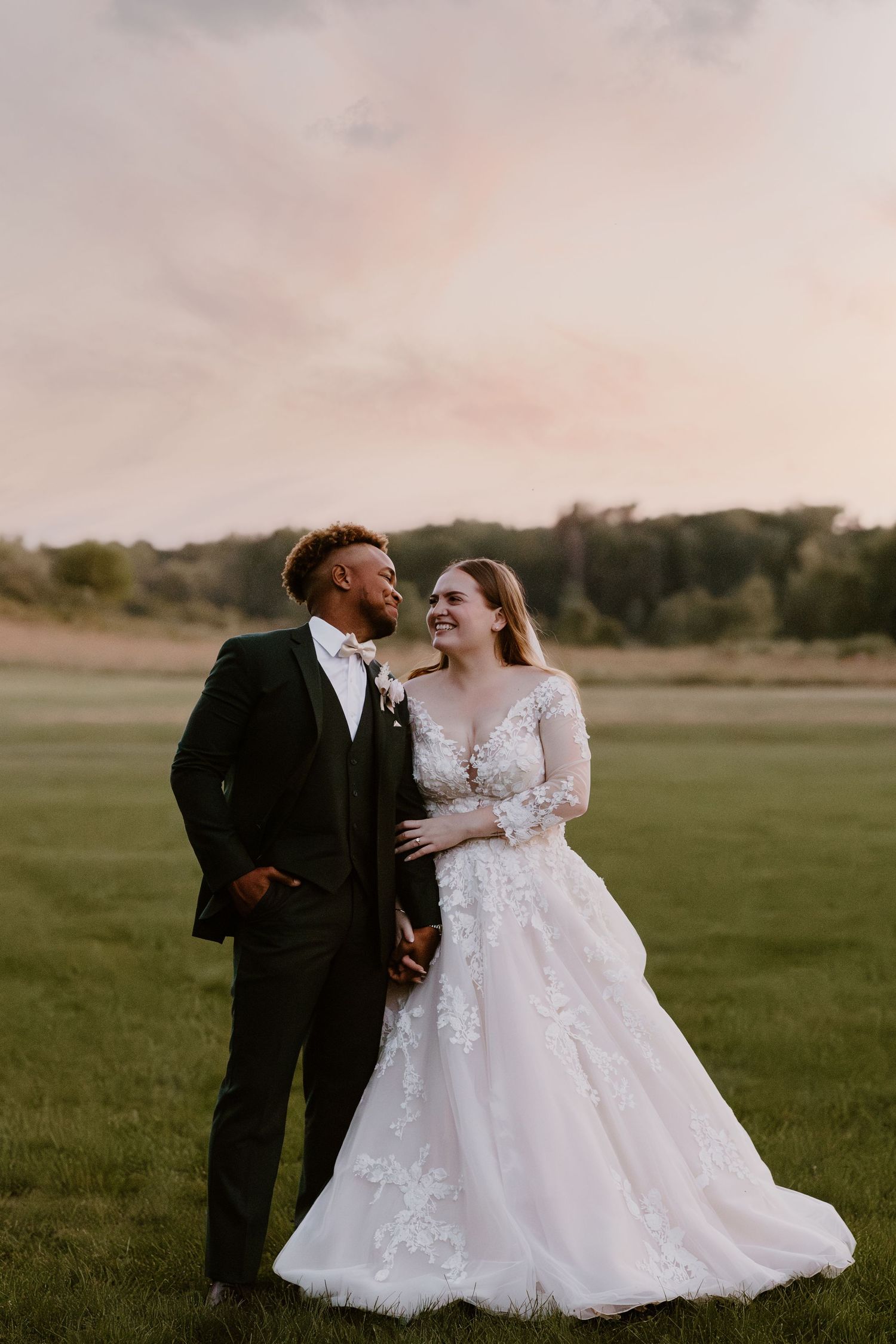 A bride and groom share a romantic moment at sunset in a lush green field, she in lace gown, he in dark tuxedo.