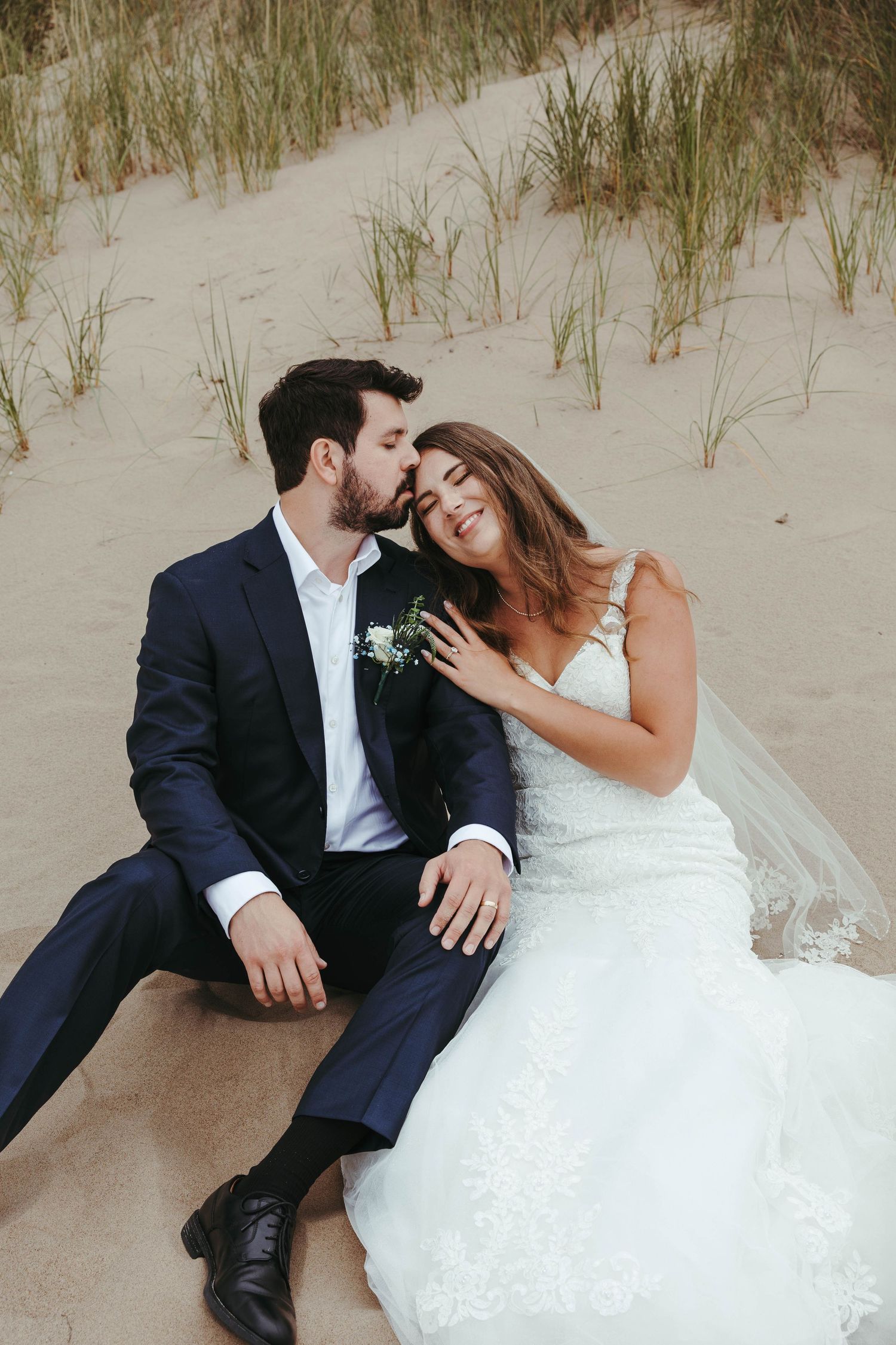 A bride and groom sit together on sandy beach dunes, sharing a romantic kiss during their wedding day.