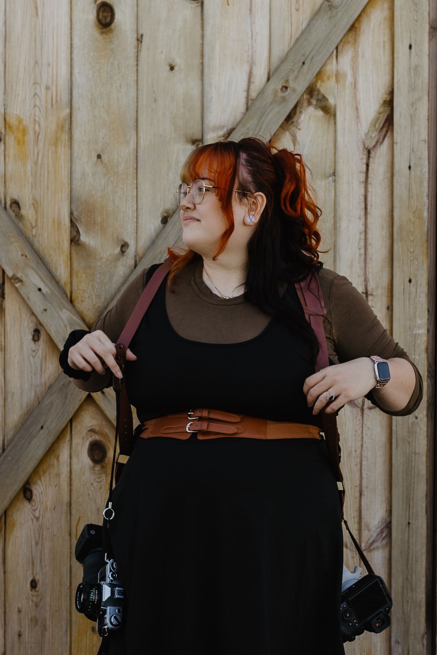 Woman with red hair wearing a black dress and brown belt posing against a rustic wooden barn door.