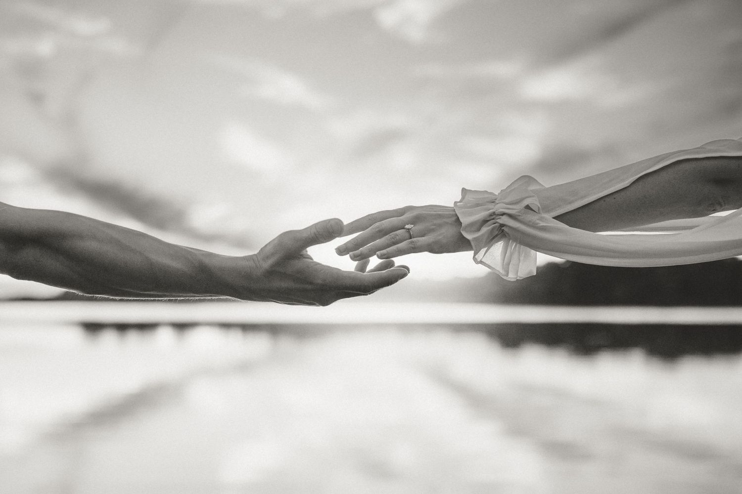 Black and white photo of two hands reaching toward each other against a cloudy sky, evoking connection and romance.