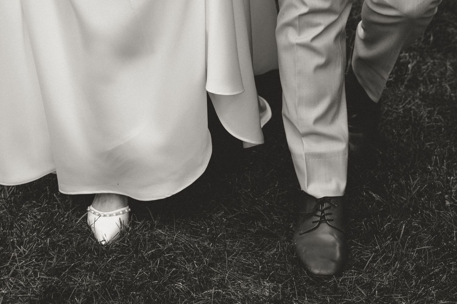 Black and white close-up of bride in white gown and groom in light suit standing on grass.
