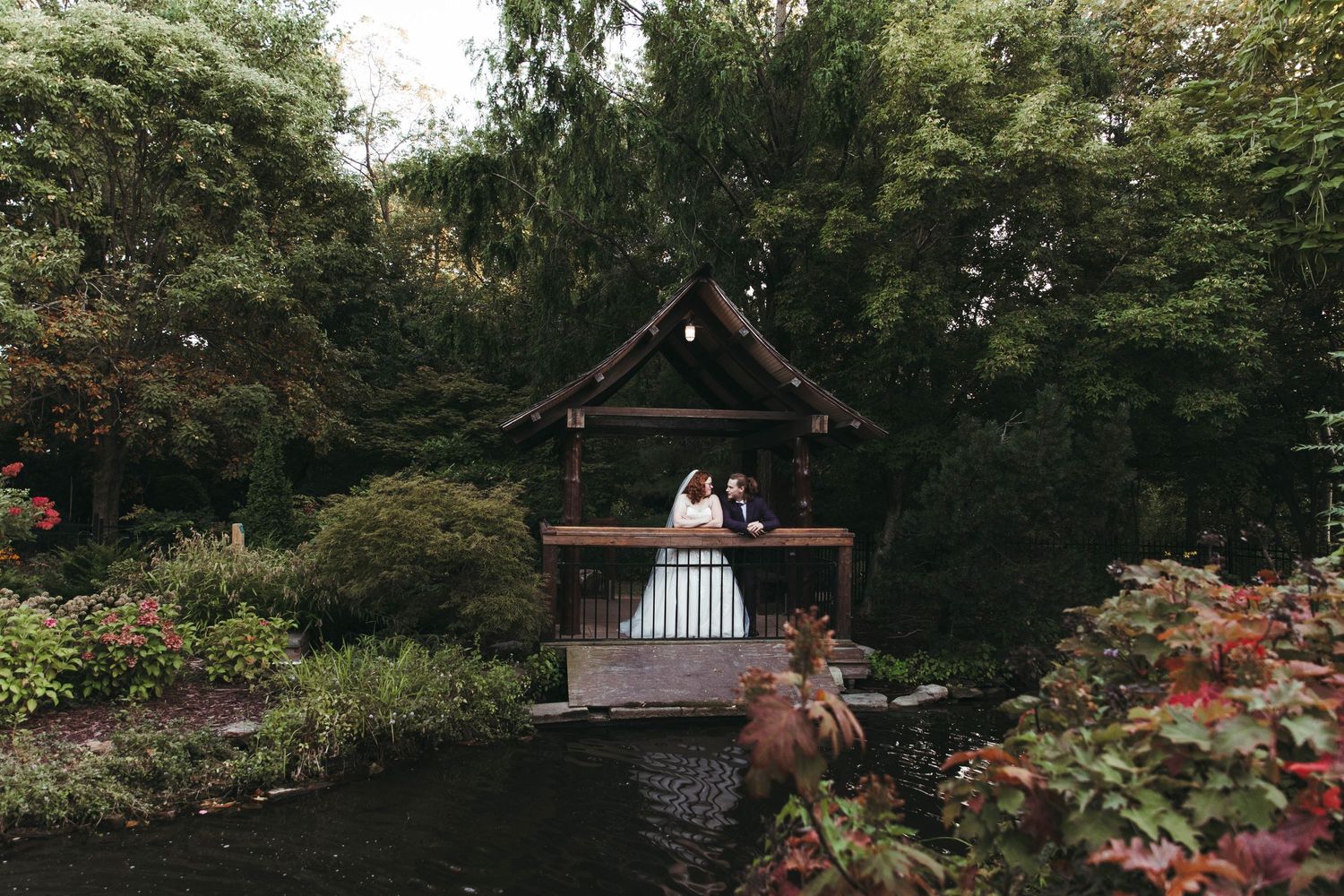 Couple in wedding attire standing on a wooden bridge gazebo surrounded by lush greenery and a serene pond.