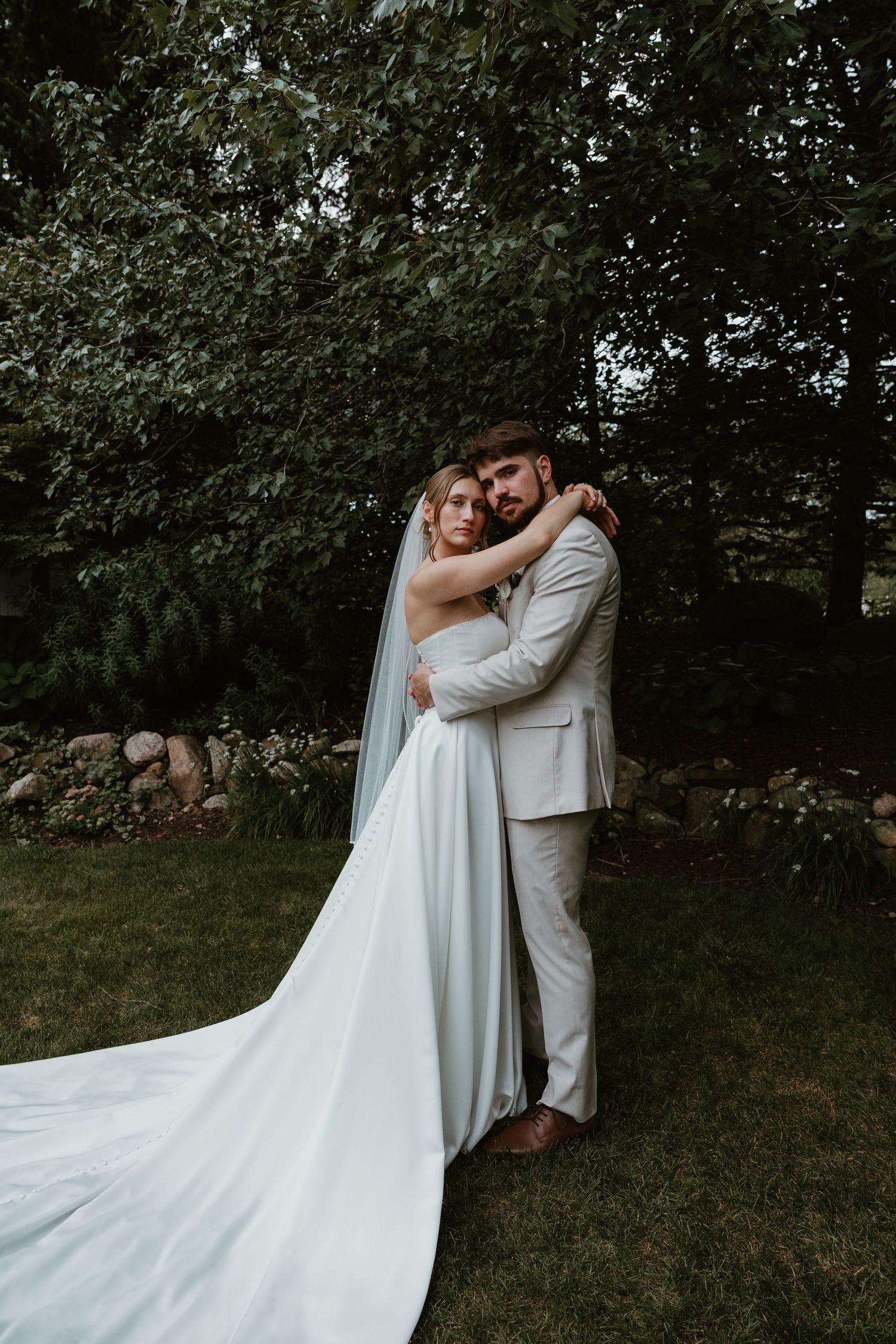 Bride and groom embracing in a lush garden, bride wearing a flowing white gown with long veil, groom in a light gray suit.