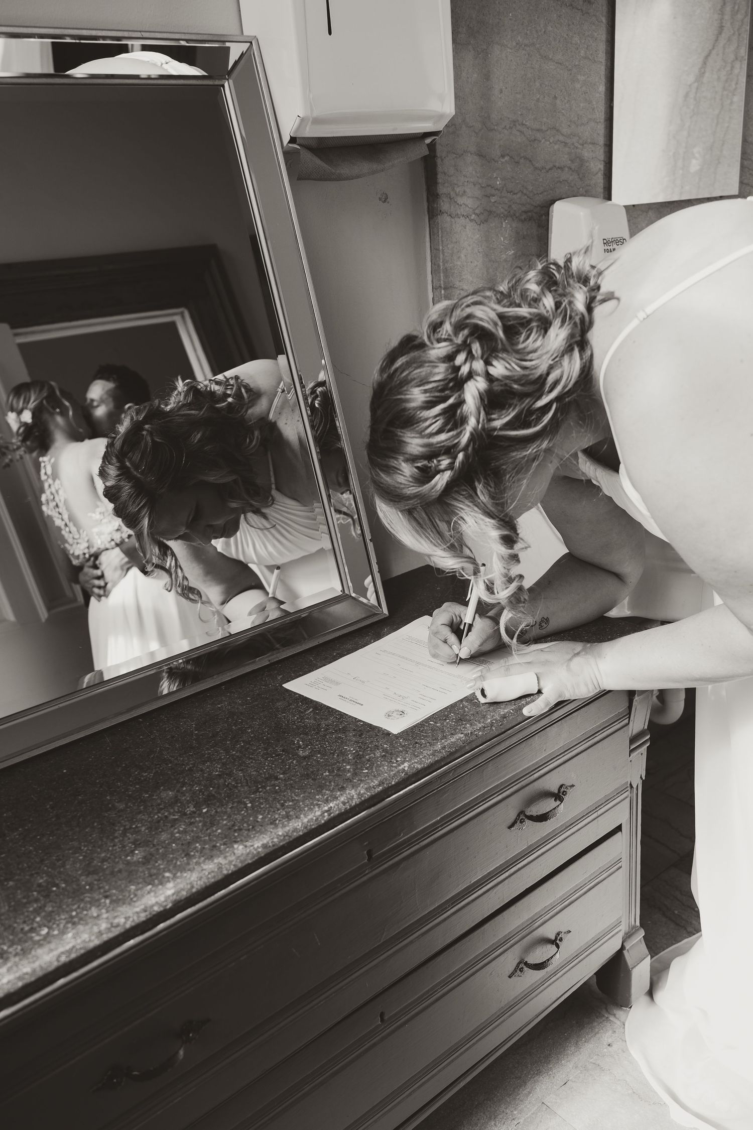 Black and white photo of bride writing vows at dresser with mirror reflection in bridal suite.