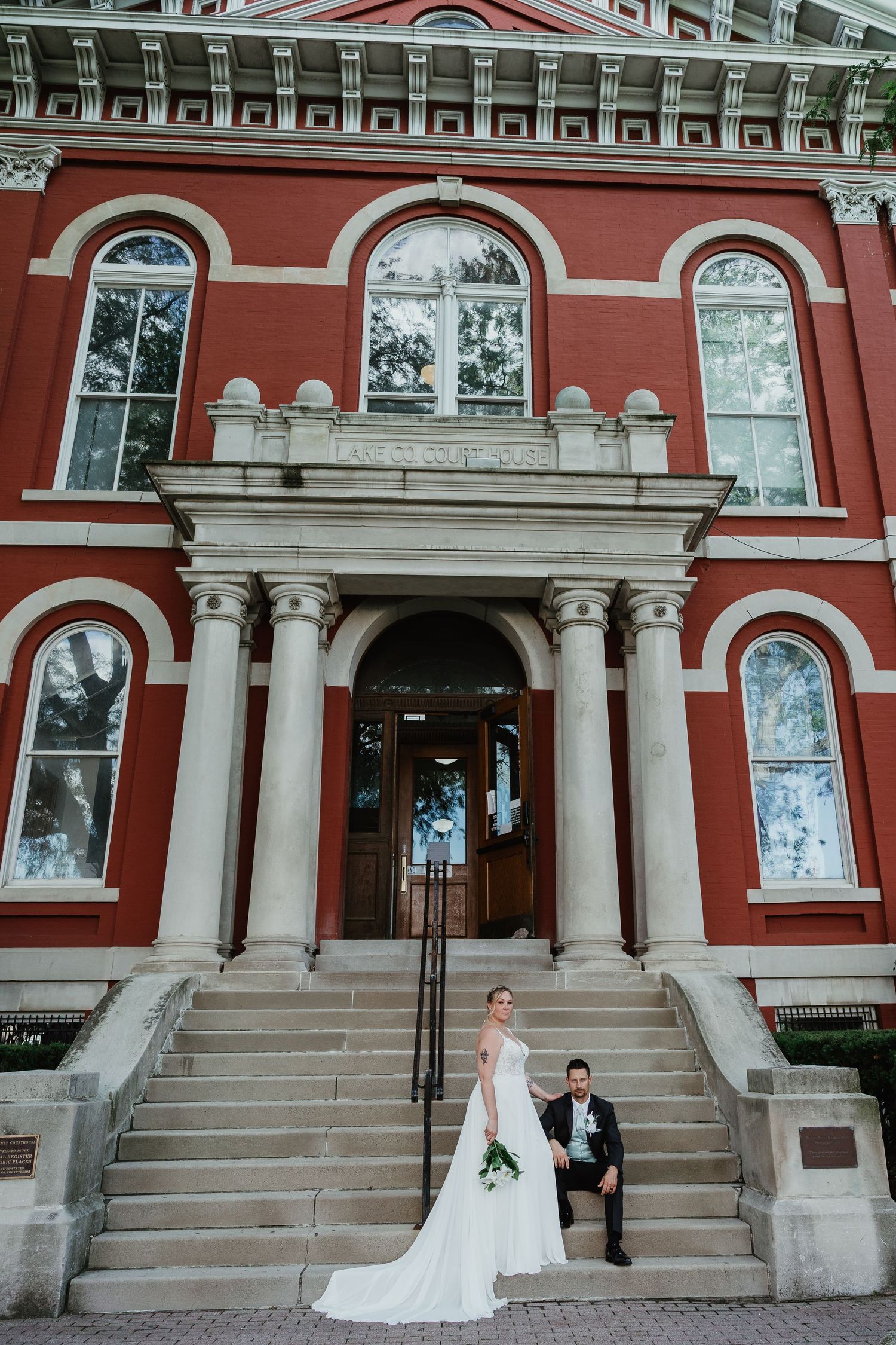 Bride and groom posing on the grand steps of a historic red brick building with white columns.