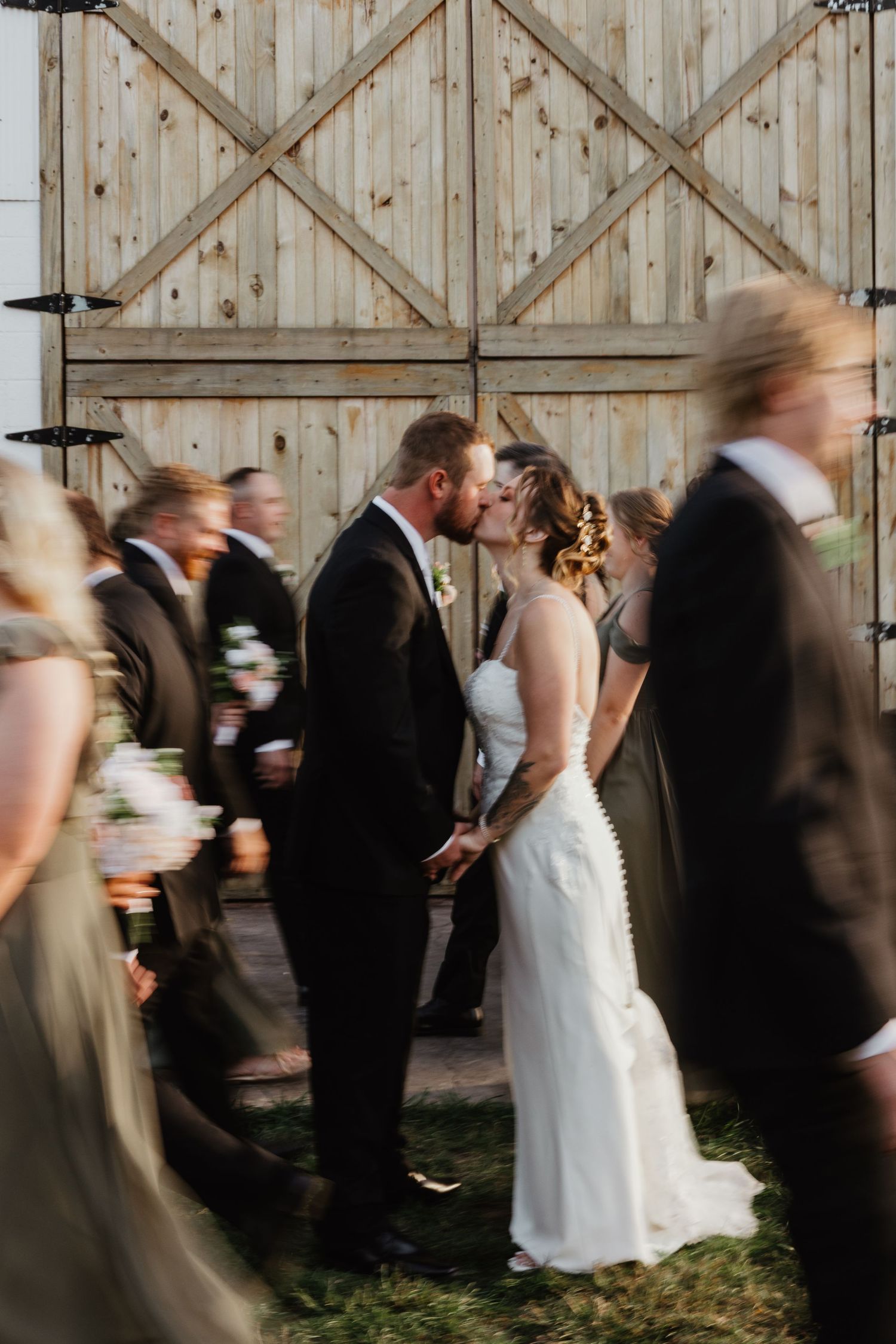 Bride and groom share first kiss at rustic barn wedding ceremony surrounded by wedding party.