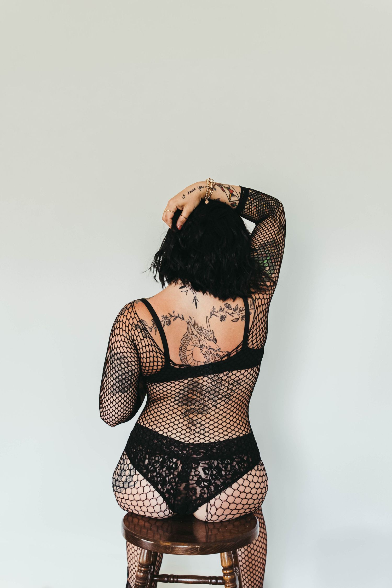 Woman in black lace lingerie and fishnet bodysuit with tattoos poses on stool showing back view.
