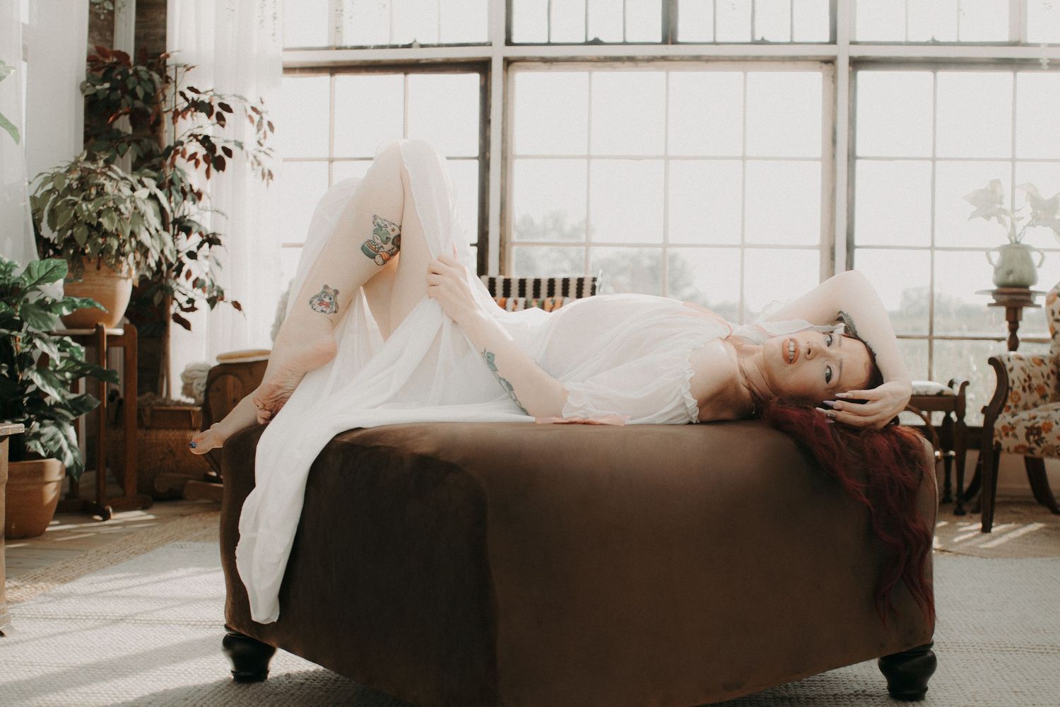 A bride in a white dress reclines on a brown chaise lounge in a sunlit loft with large windows and plants.