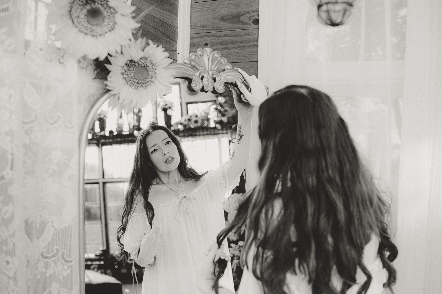 Black and white photo of a young woman with long curly hair looking at her reflection in an ornate mirror.