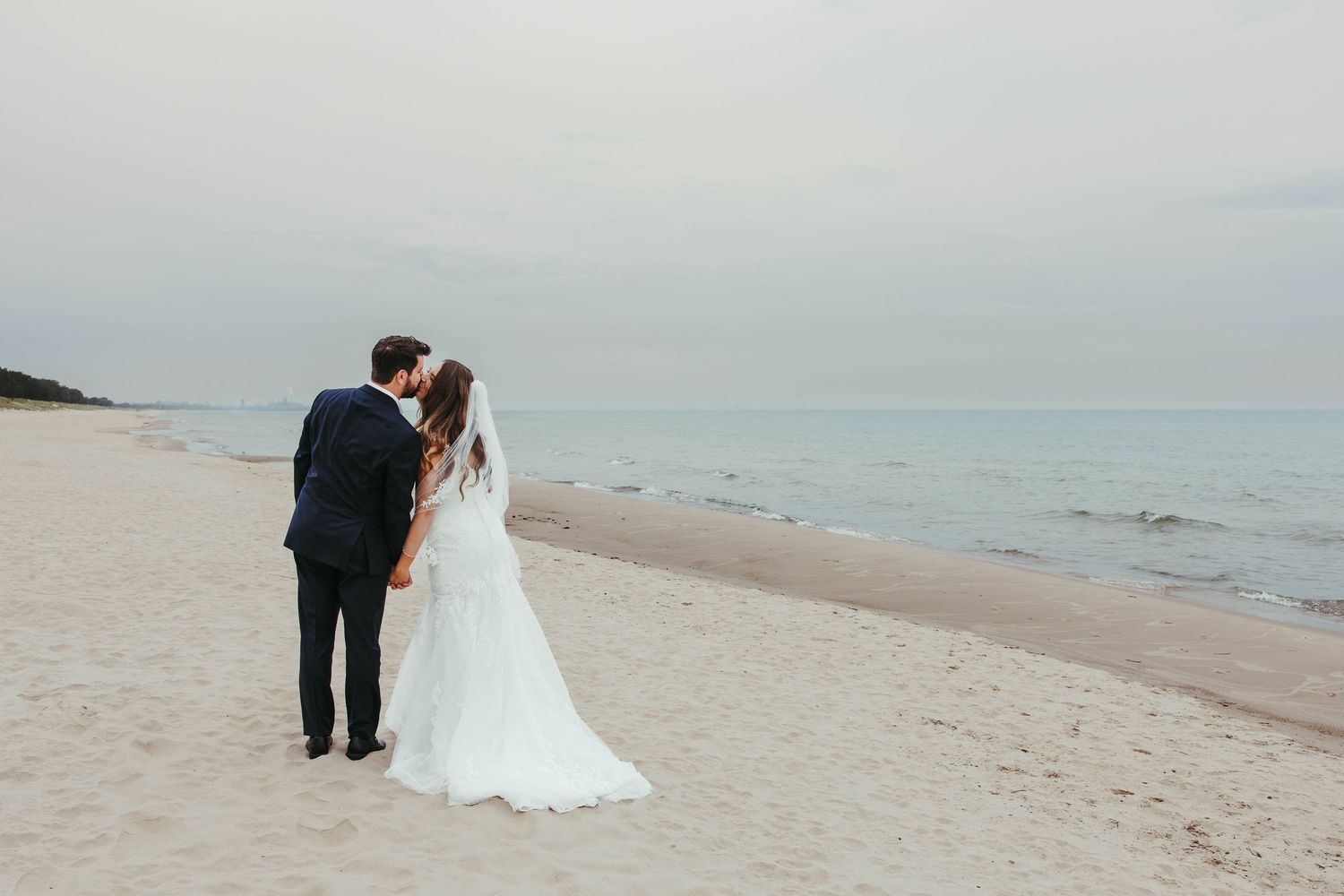 Bride and groom sharing a romantic kiss on a serene sandy beach at dusk.