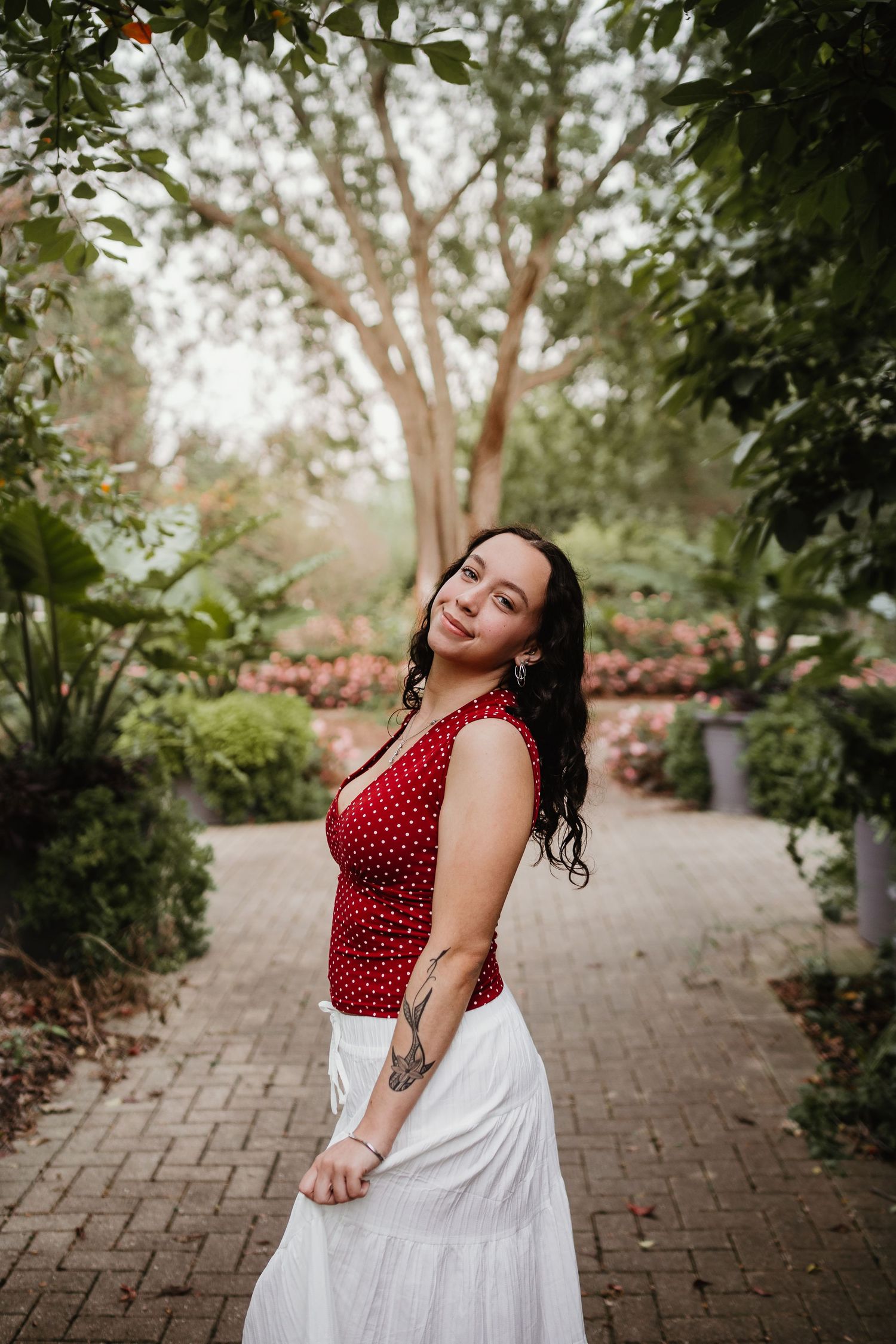 Young woman in red polka dot top and white skirt posing in a lush green garden pathway.