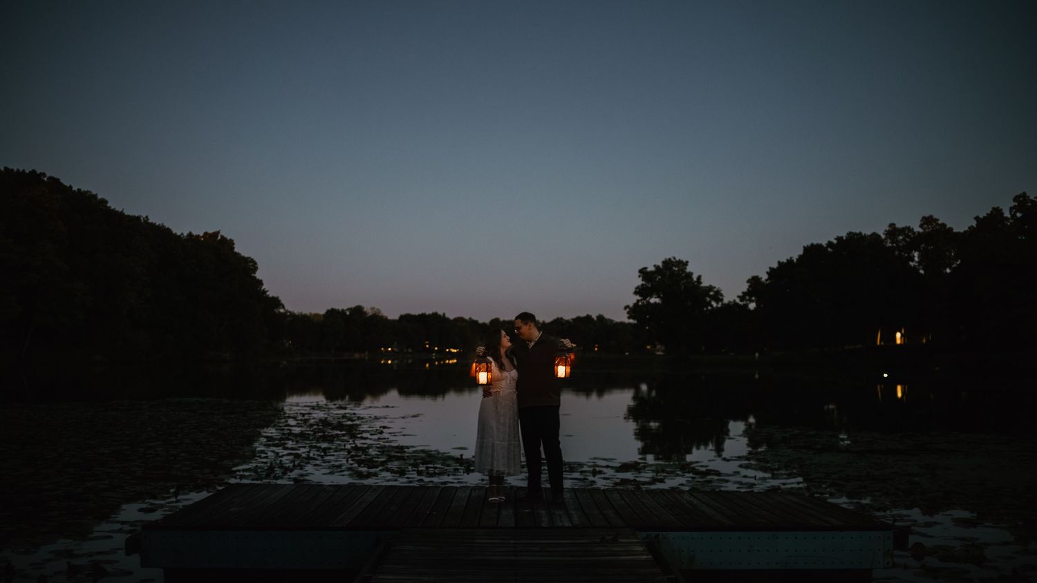Couple holding lanterns by a tranquil pond at dusk, creating a romantic silhouette against the twilight sky.