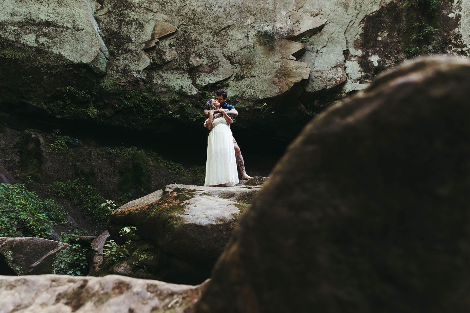 Couple embracing romantically in a cave entrance surrounded by rocks and lush greenery in a scenic outdoor setting.