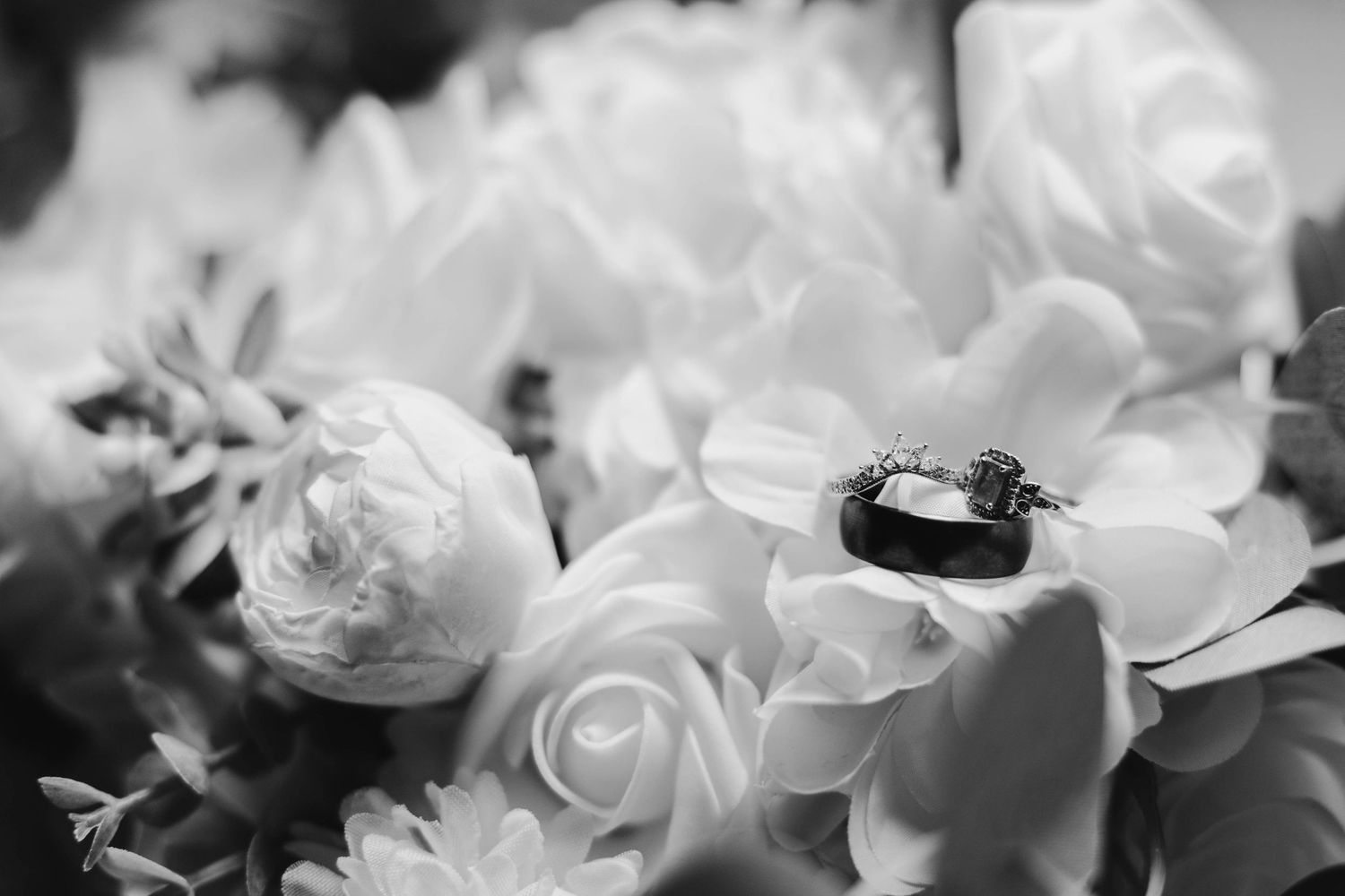 Black and white photo of wedding rings resting on a bouquet of white roses and flowers.