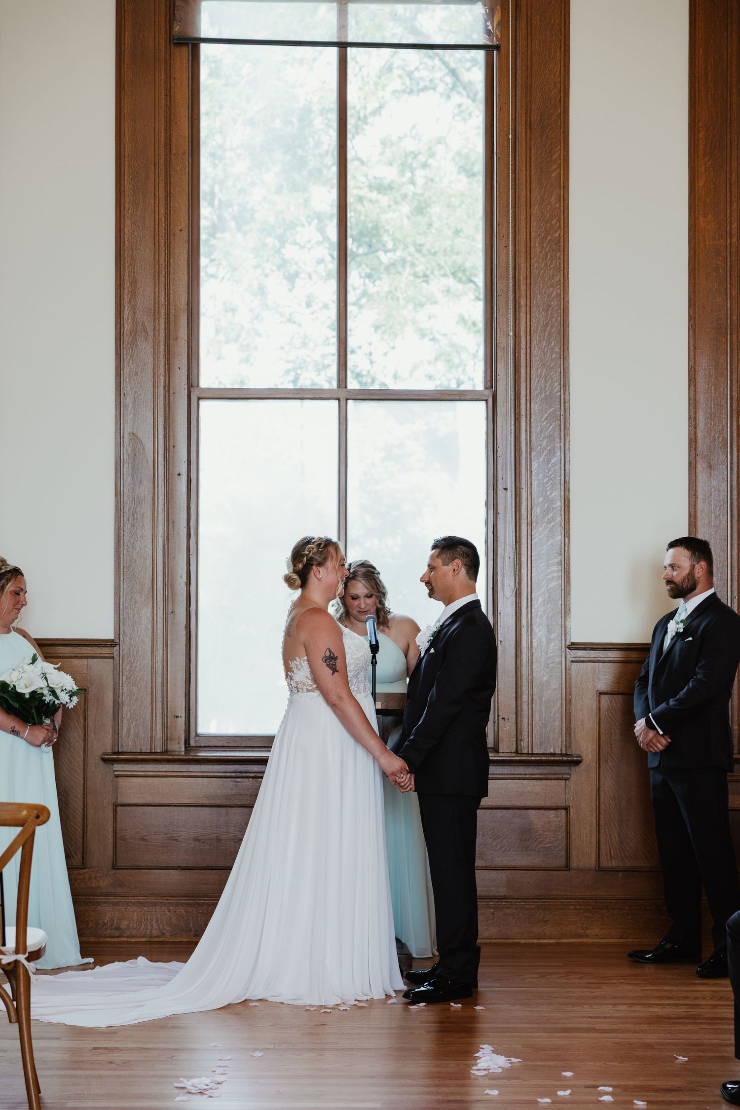 Bride and groom exchange vows at indoor wedding ceremony before large wood-framed window with officiant and wedding party.